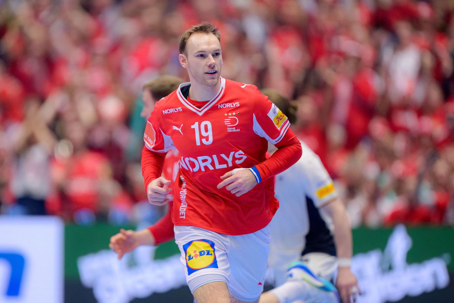 Handball player wearing red jersey number 19 with Norlys sponsor sprinting on indoor court.