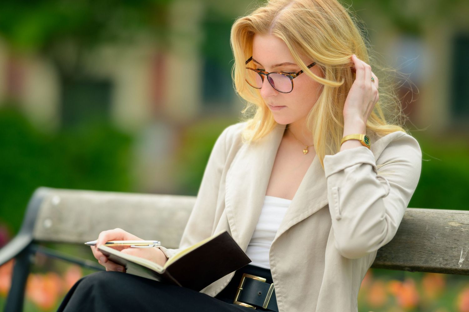 Someone wearing glasses sits on a park bench while reading a book on a sunny day, tucking blonde hair behind their ear.
