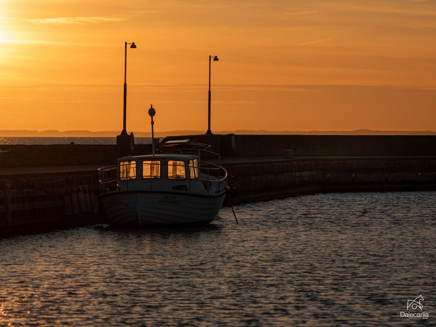 A white boat moored at a harbor pier during a stunning golden sunset over calm waters.