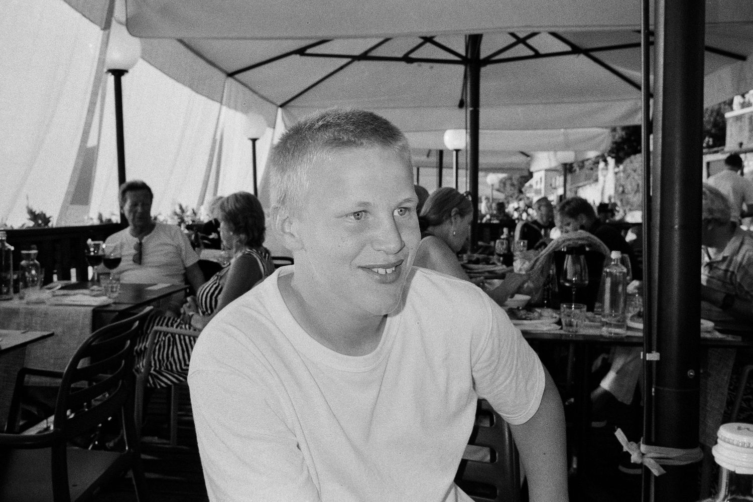 A person in a white t-shirt sits smiling at an outdoor cafe or restaurant patio under umbrellas.