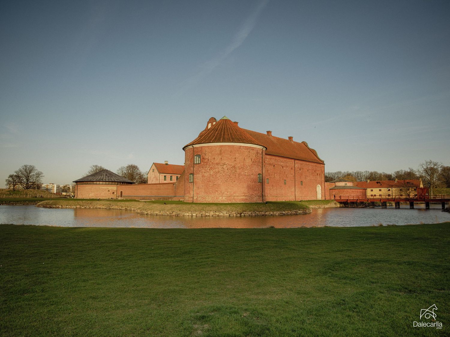 Historic red brick castle surrounded by a moat with green lawns under a clear blue sky at golden hour.