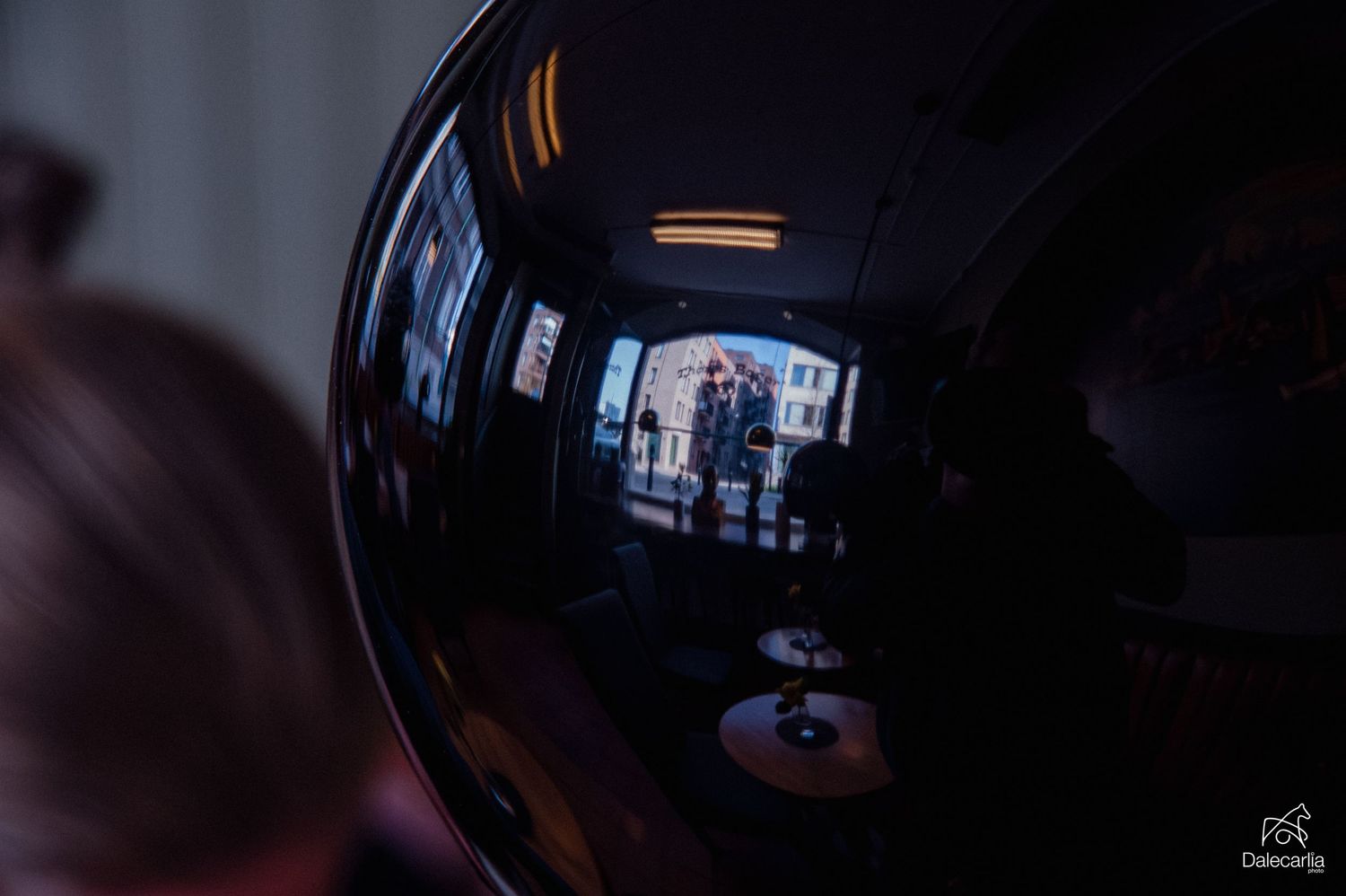A reflective black sphere captures distorted mirror image of two people standing outdoors in a dimly lit interior setting.
