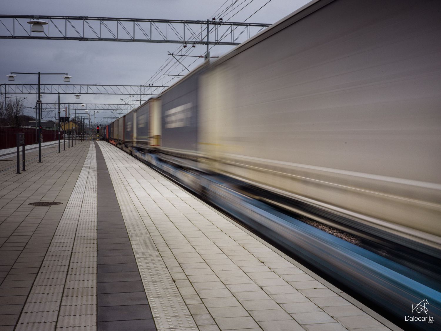 A blurred train speeds past an empty railway platform with overhead power lines and tiled flooring.