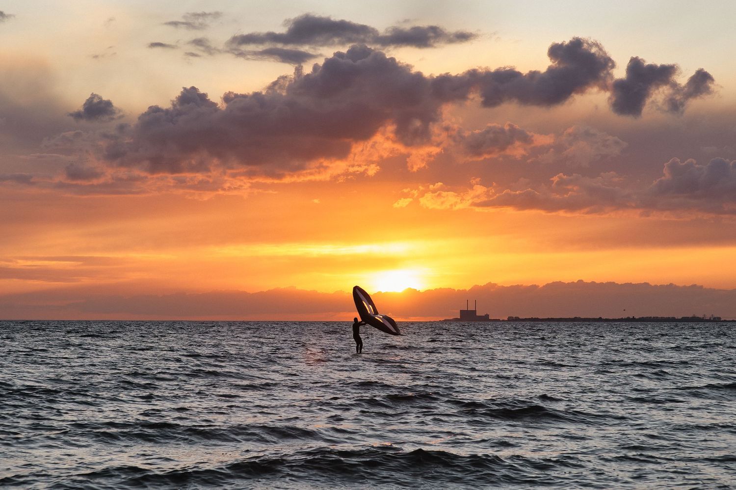 A silhouetted surfer catches a wave at sunset with vibrant orange clouds and sun reflecting on the ocean waters.