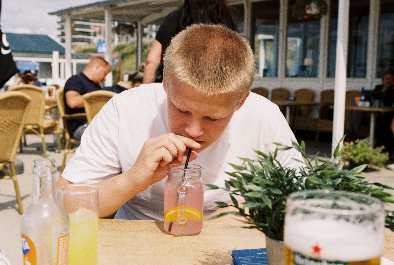 Person in white t-shirt sips a drink through a straw at an outdoor restaurant patio with beverages on the table.