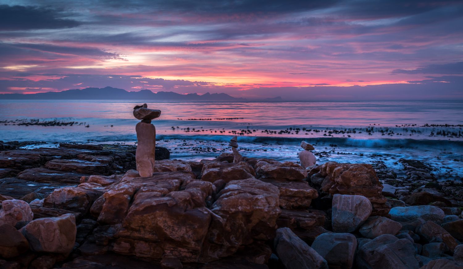 Stunning coastal sunset with vibrant pink and purple sky, rocky shoreline, and stacked stone cairns overlooking calm ocean waters.