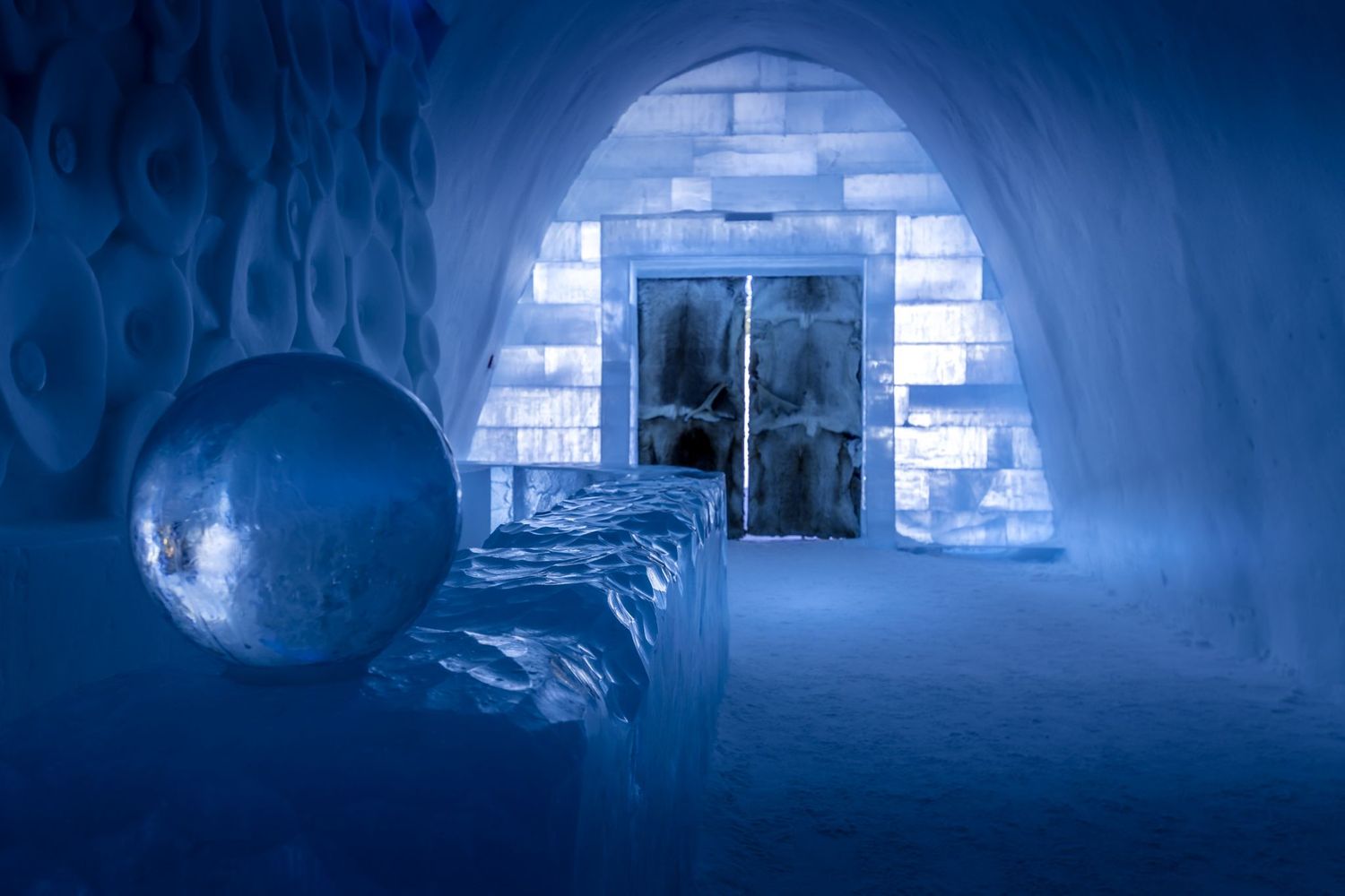 Ice hotel interior with glowing blue walls, arched ceiling, silver orb sculpture, and illuminated ice block doorway.