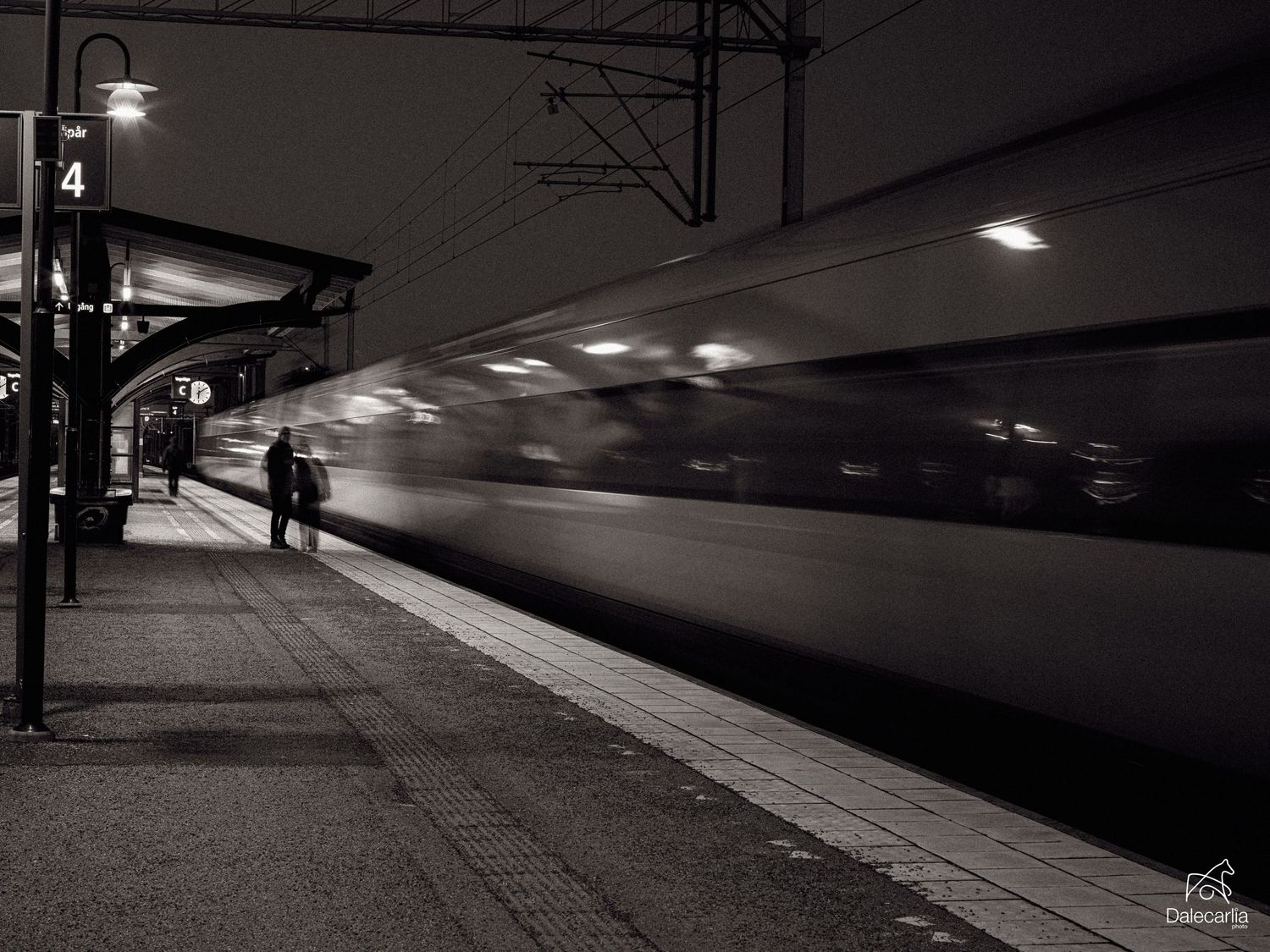 A dark train platform at night illuminated by overhead lights casting shadows across the empty station.