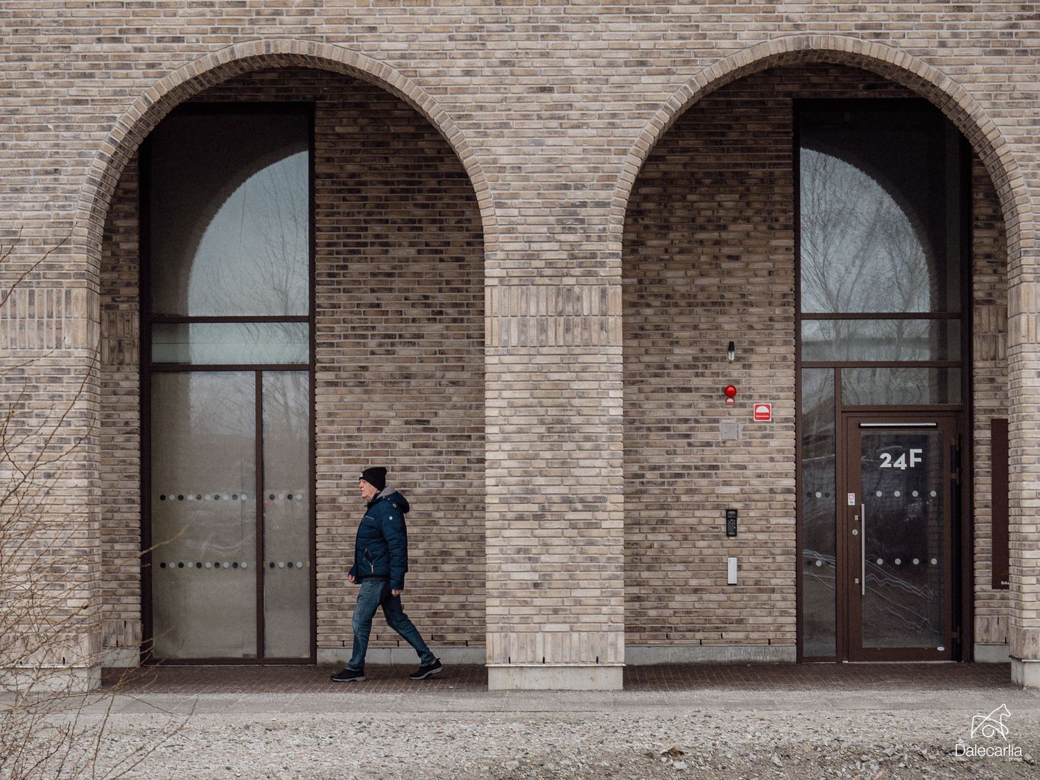 A person in blue walks past tall arched doorways in a modern brick building facade.
