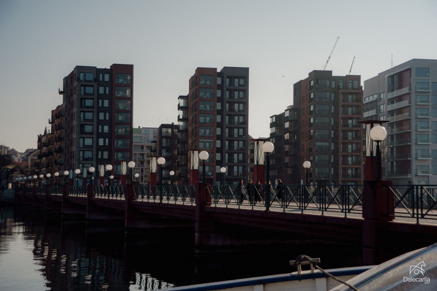 Modern waterfront apartment buildings with red railings on a pedestrian bridge reflected in calm harbor waters at dusk.