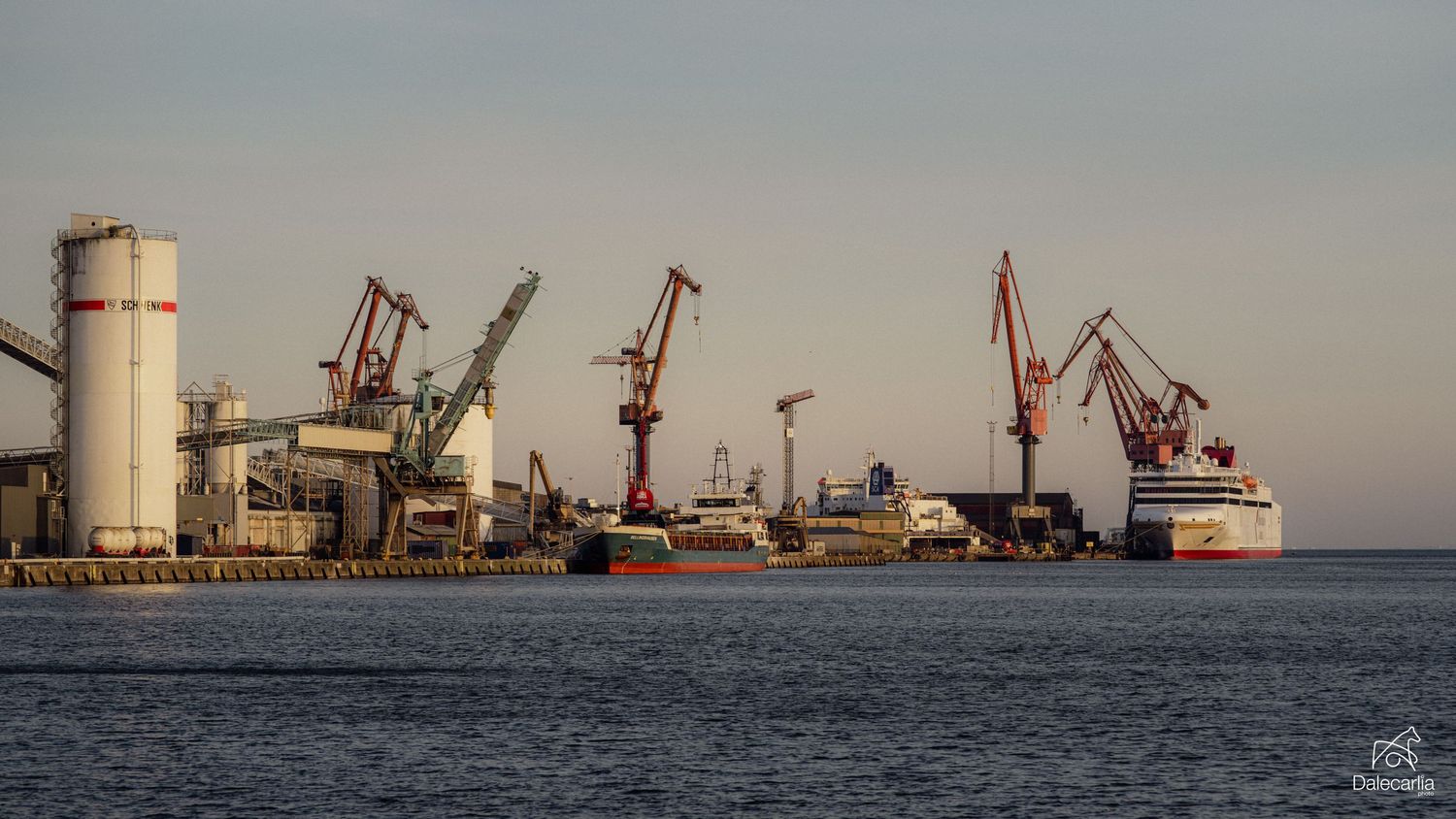 Industrial harbor with red cranes, cargo ships, and a cruise liner docked at sunset on calm blue waters.