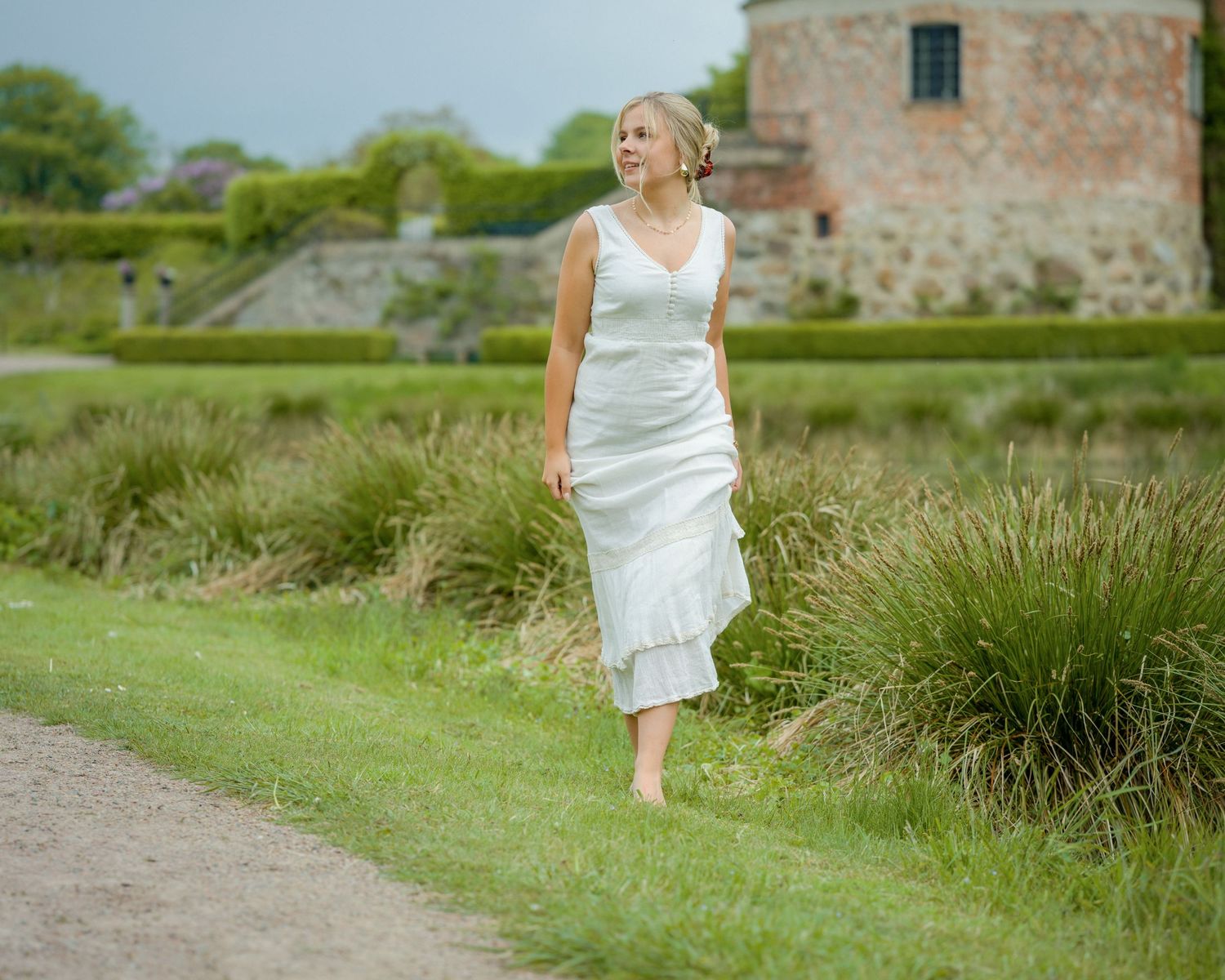 A person in a flowing white dress walks along a countryside path with an old stone building in the background.