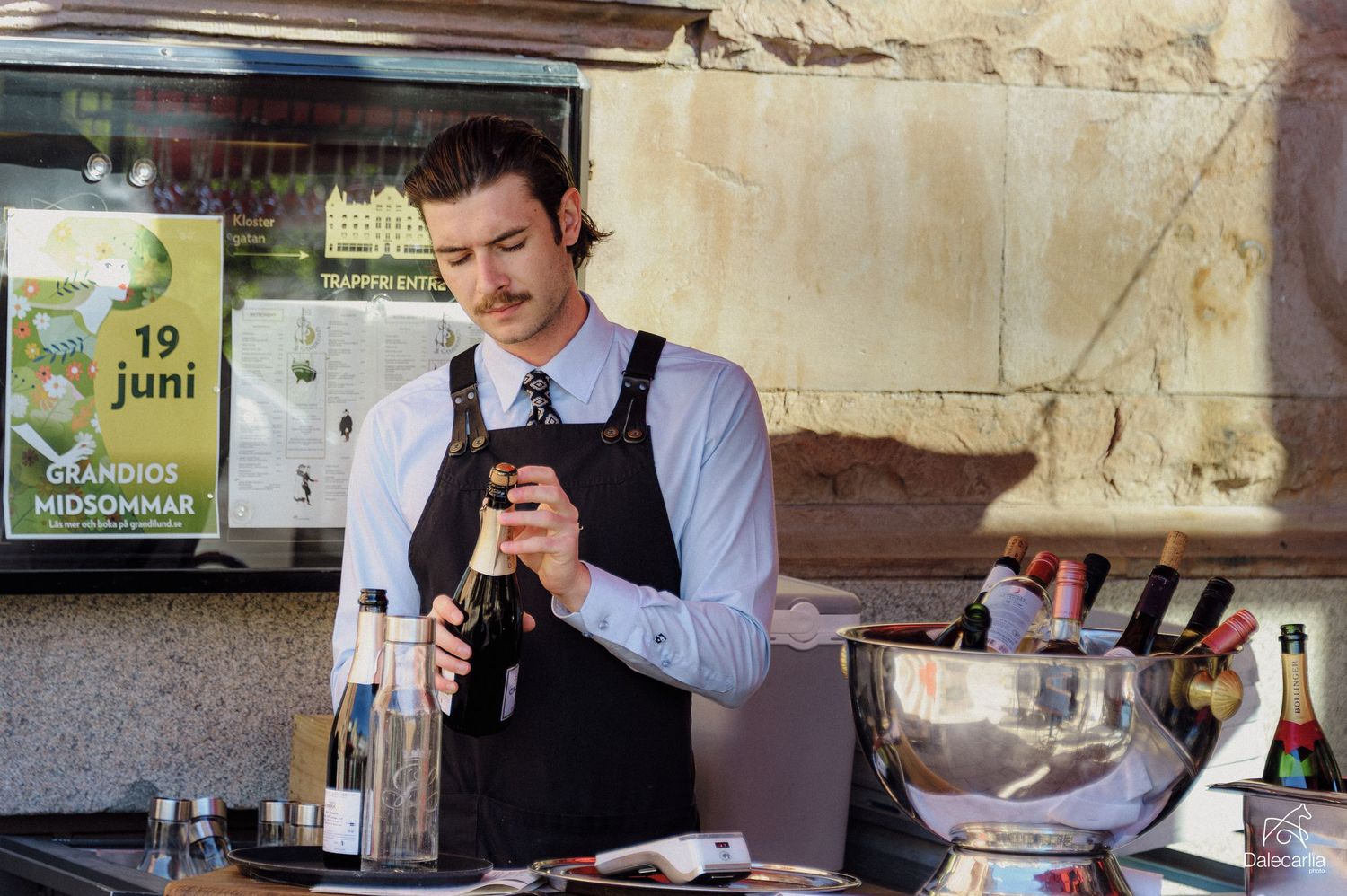 A bartender in a black apron opens a wine bottle at an outdoor bar with bottles and an ice bucket nearby.