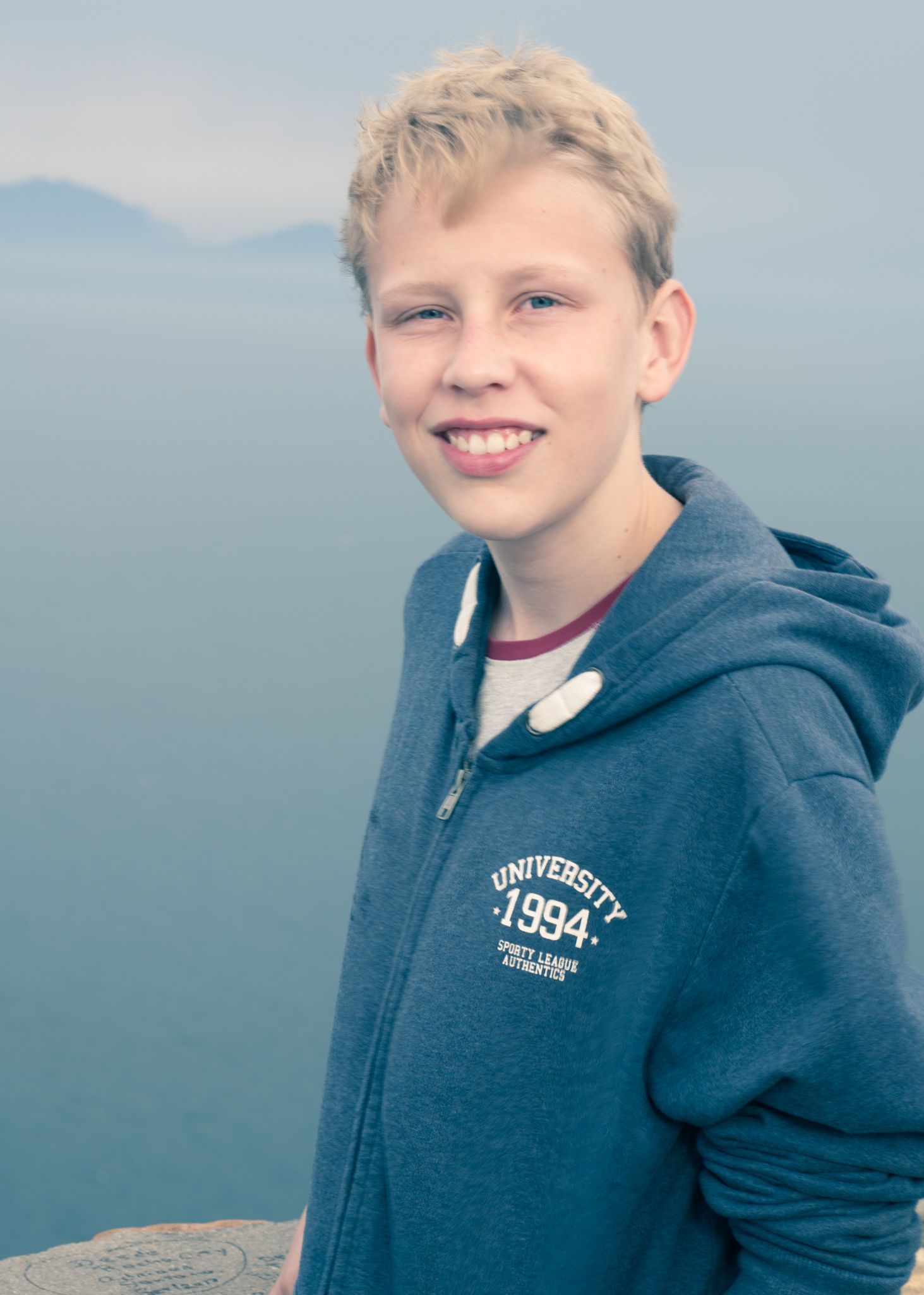 Smiling teenage boy with blonde hair wearing a blue University 1994 hoodie outdoors with misty mountains in background.
