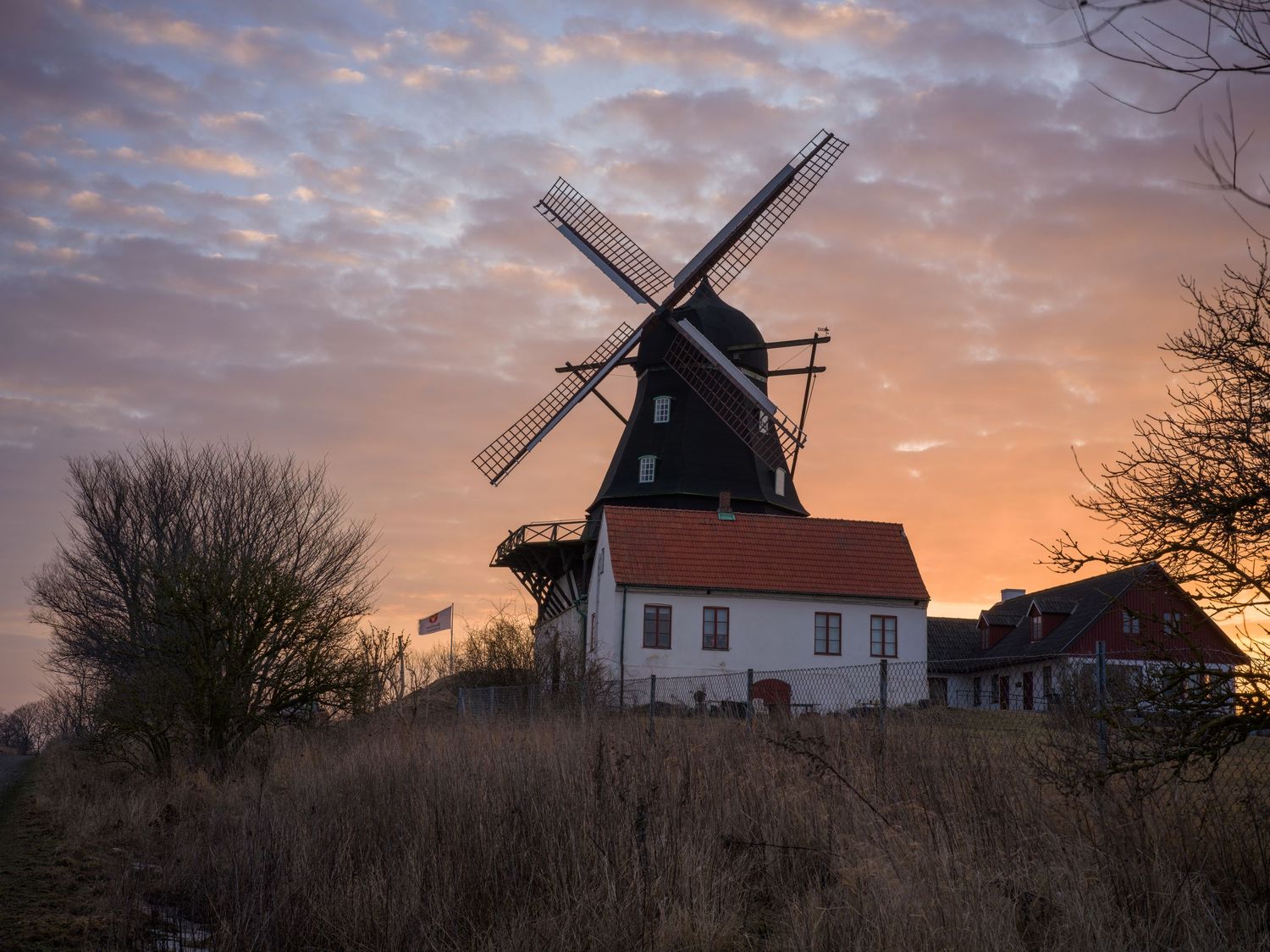 Traditional Dutch windmill silhouetted against a dramatic sunset sky with pink clouds and a white building below.