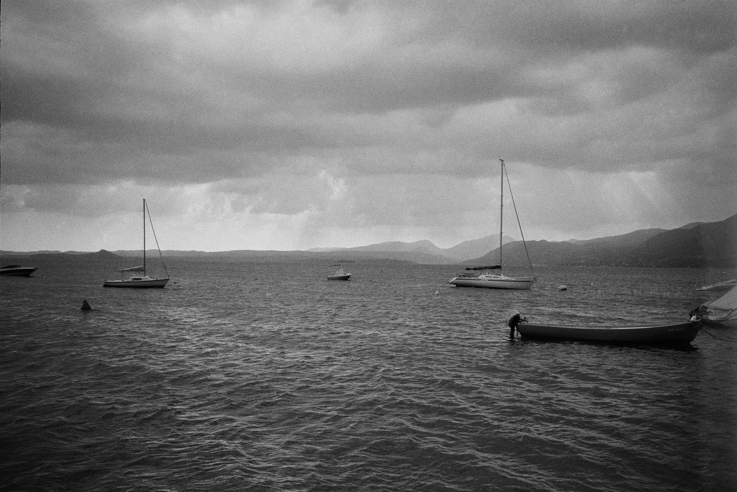 Sailboats float serenely on calm waters against a moody black and white cloudy sky and mountain backdrop.