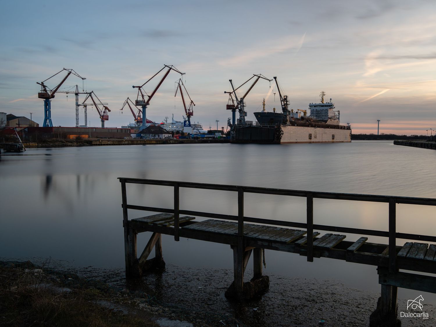 Industrial harbor at dusk with large cranes and cargo ship reflected in calm water, viewed from a wooden dock.