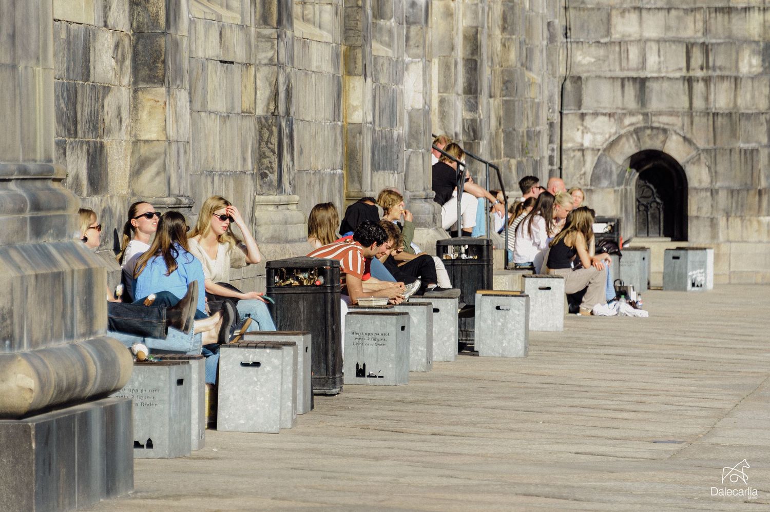 People relaxing on stone benches along a historic castle wall on a sunny day.