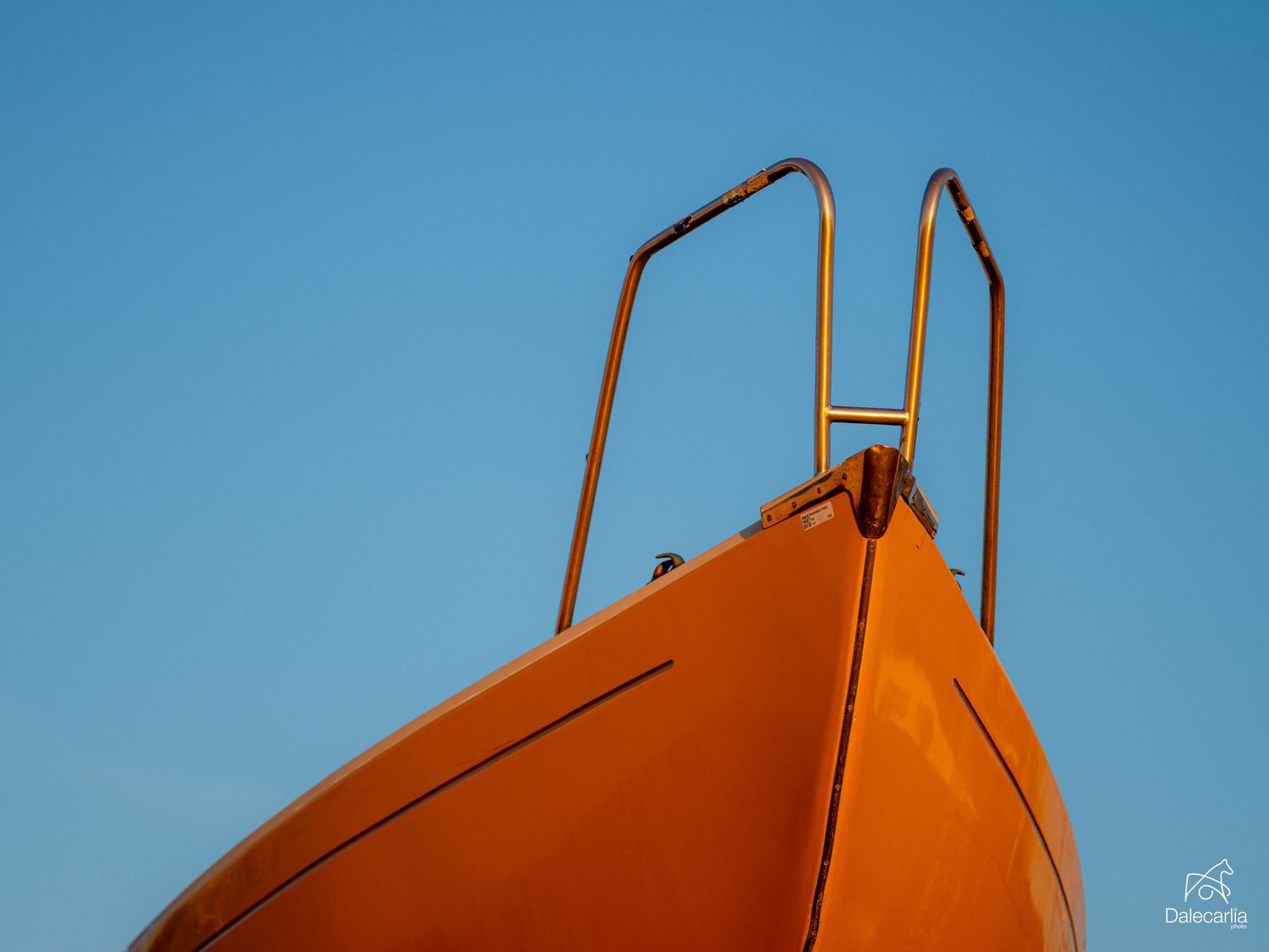 Close-up of a bright orange boat bow against a clear blue sky, featuring shiny metal railings on top.