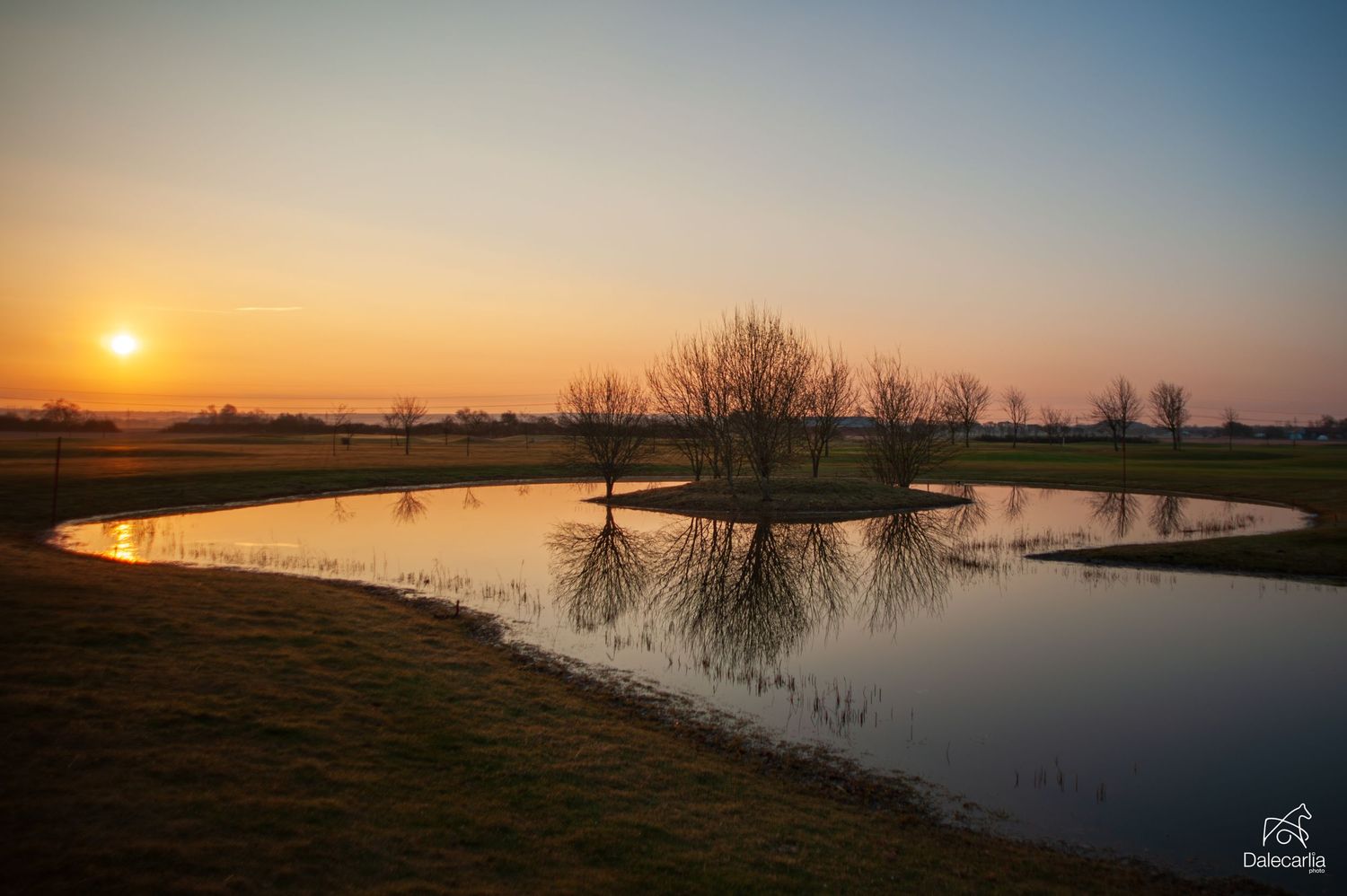 Serene sunset reflecting on a calm pond with bare trees silhouetted against an orange and blue sky.