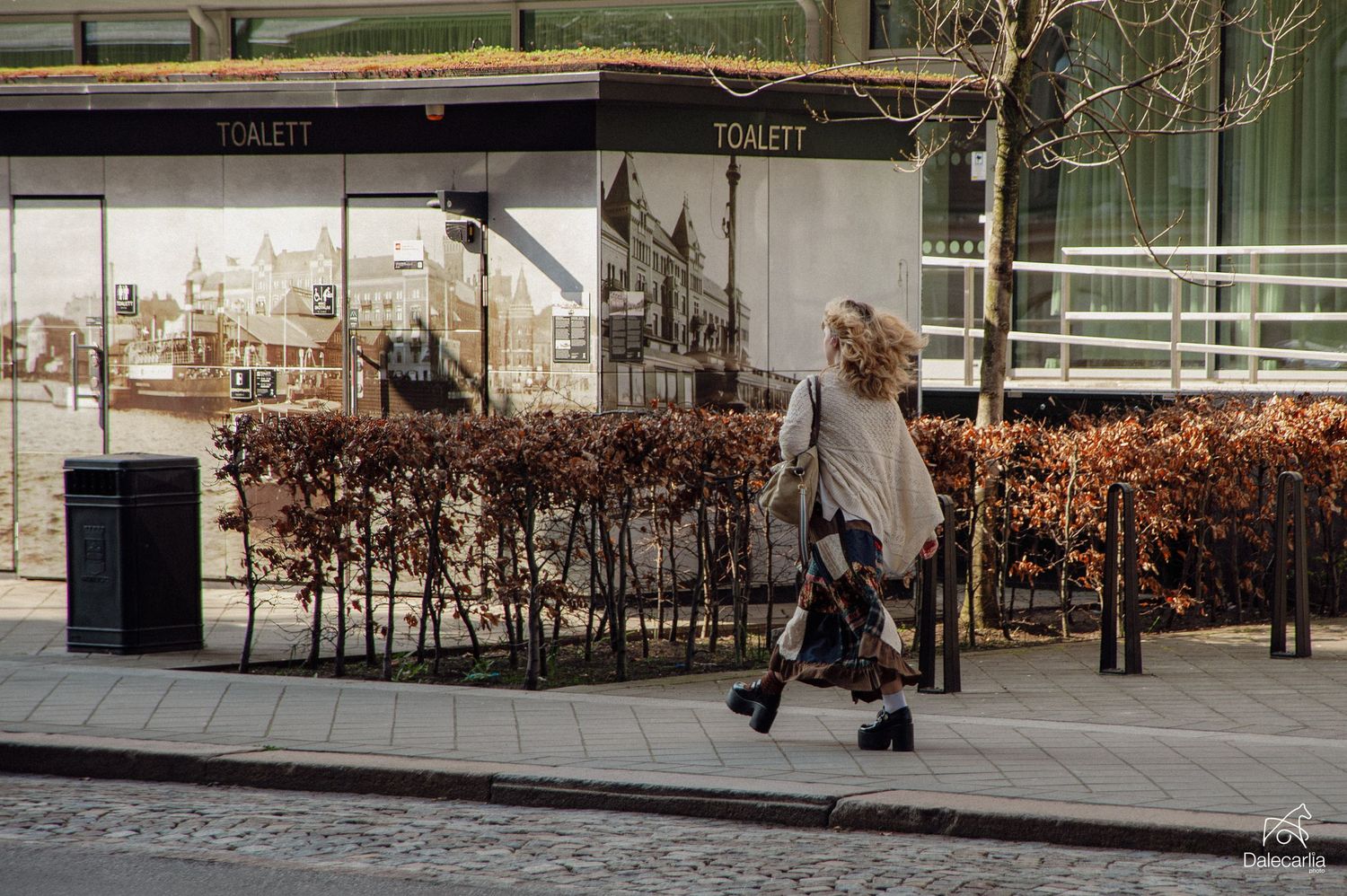 Elderly woman with walker strolling past a public toilet building with autumn bushes on a city sidewalk.