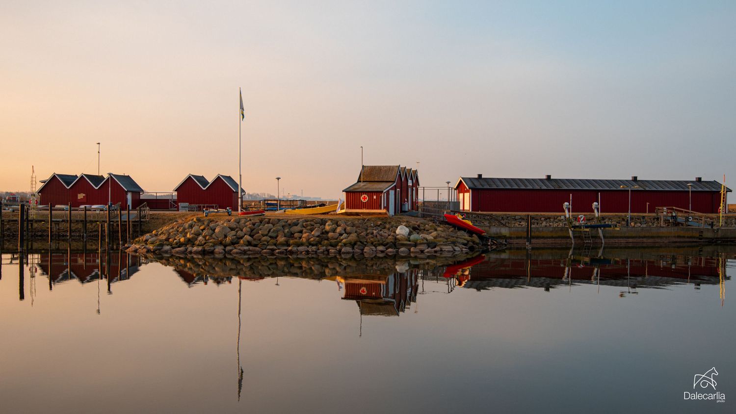 A panoramic view of boats and houseboats docked at a marina during sunset with orange sky reflecting on calm water.