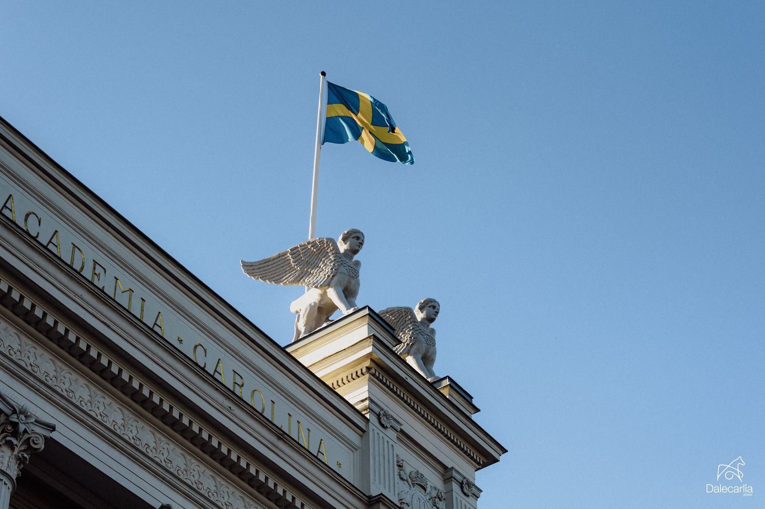 Swedish flag waving atop a historic building with stone sculptures against a clear blue sky.