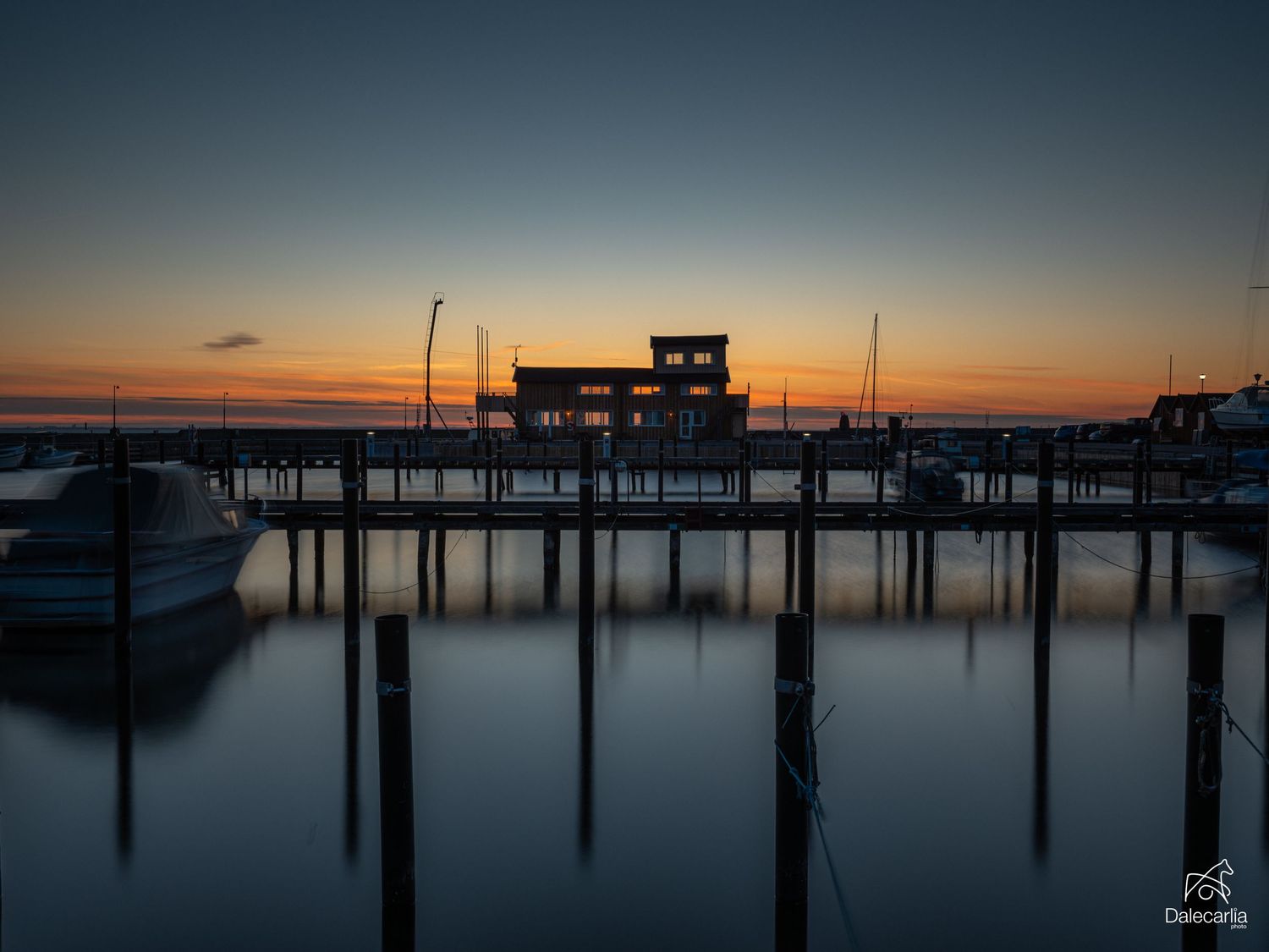 Serene marina at sunset with silhouetted dock building, boats, and pier pilings reflected in calm water.