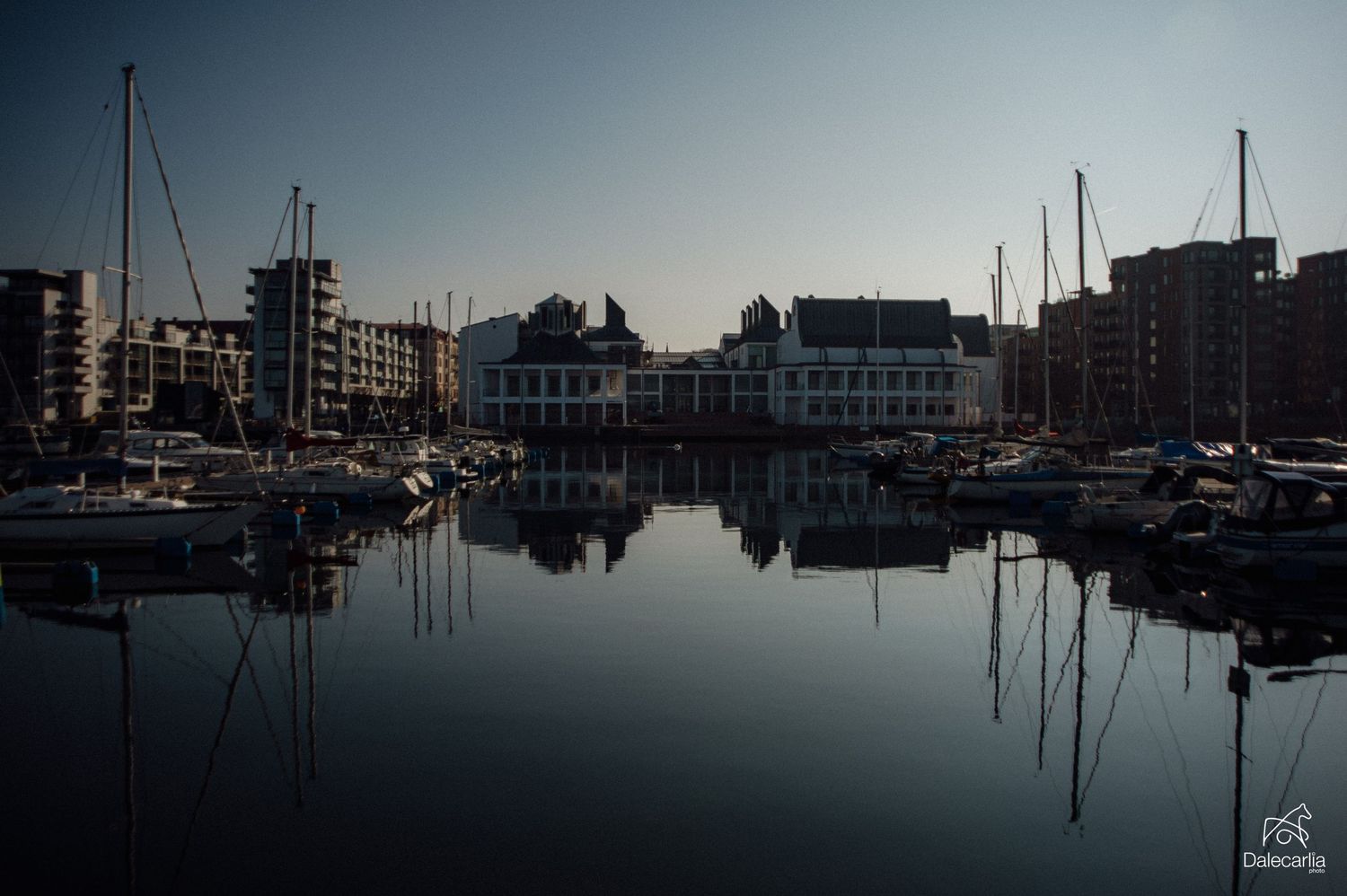 Calm marina at dusk with sailboats and yachts reflecting on still water, framed by waterfront buildings.