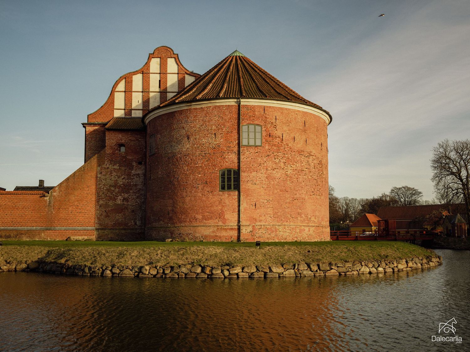 Historic red brick castle tower with conical roof reflected in surrounding moat under golden evening light.
