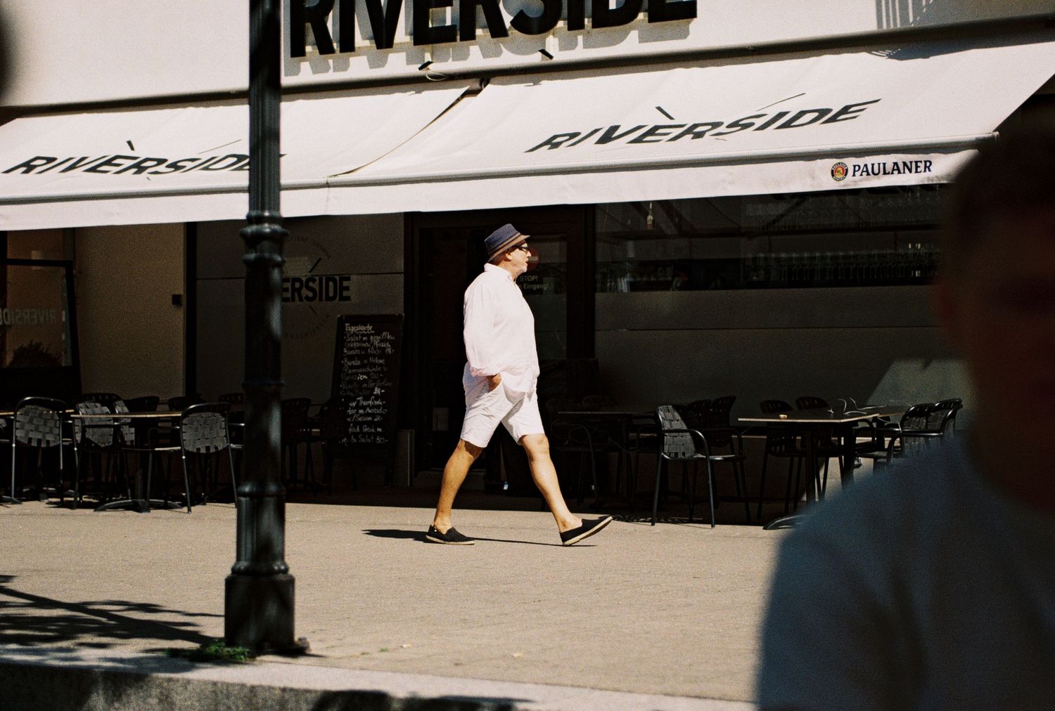 A figure in all white clothing walks past a Riverside storefront on a sunny day.