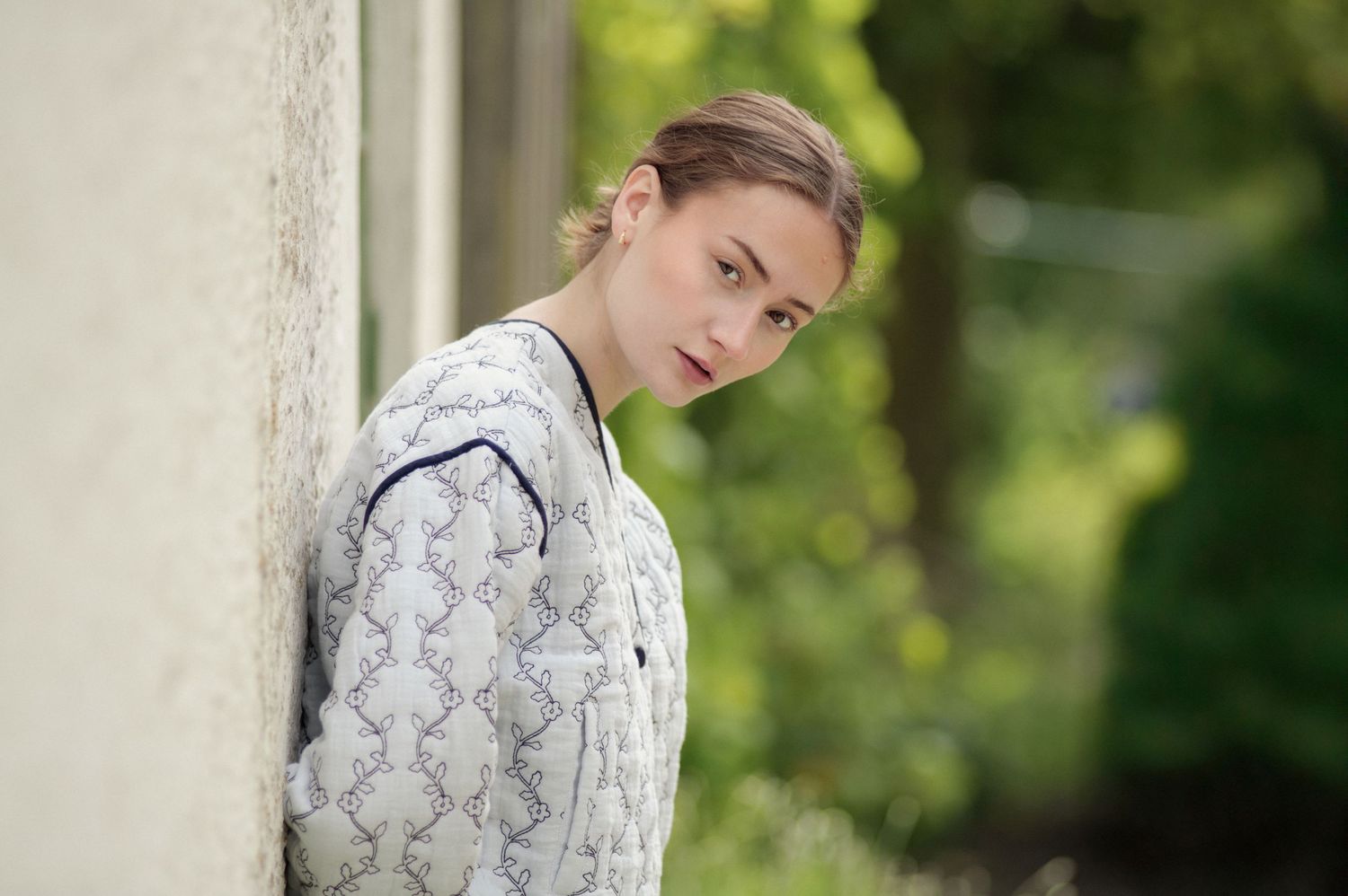 Person in white patterned sweater leans against a wall outdoors with blurred green foliage in background.