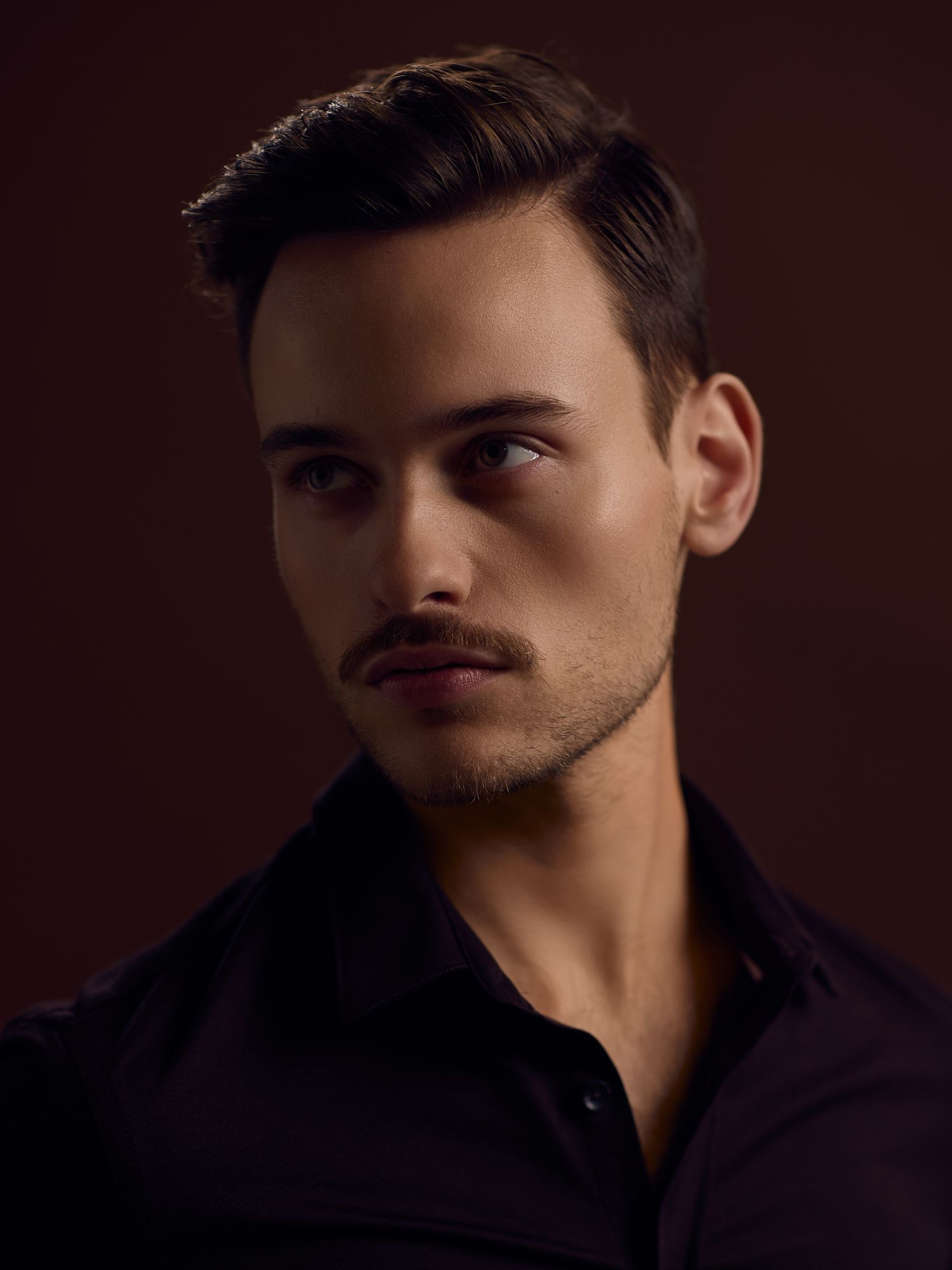 Moody portrait series in dark lighting wearing black shirt against burgundy background.