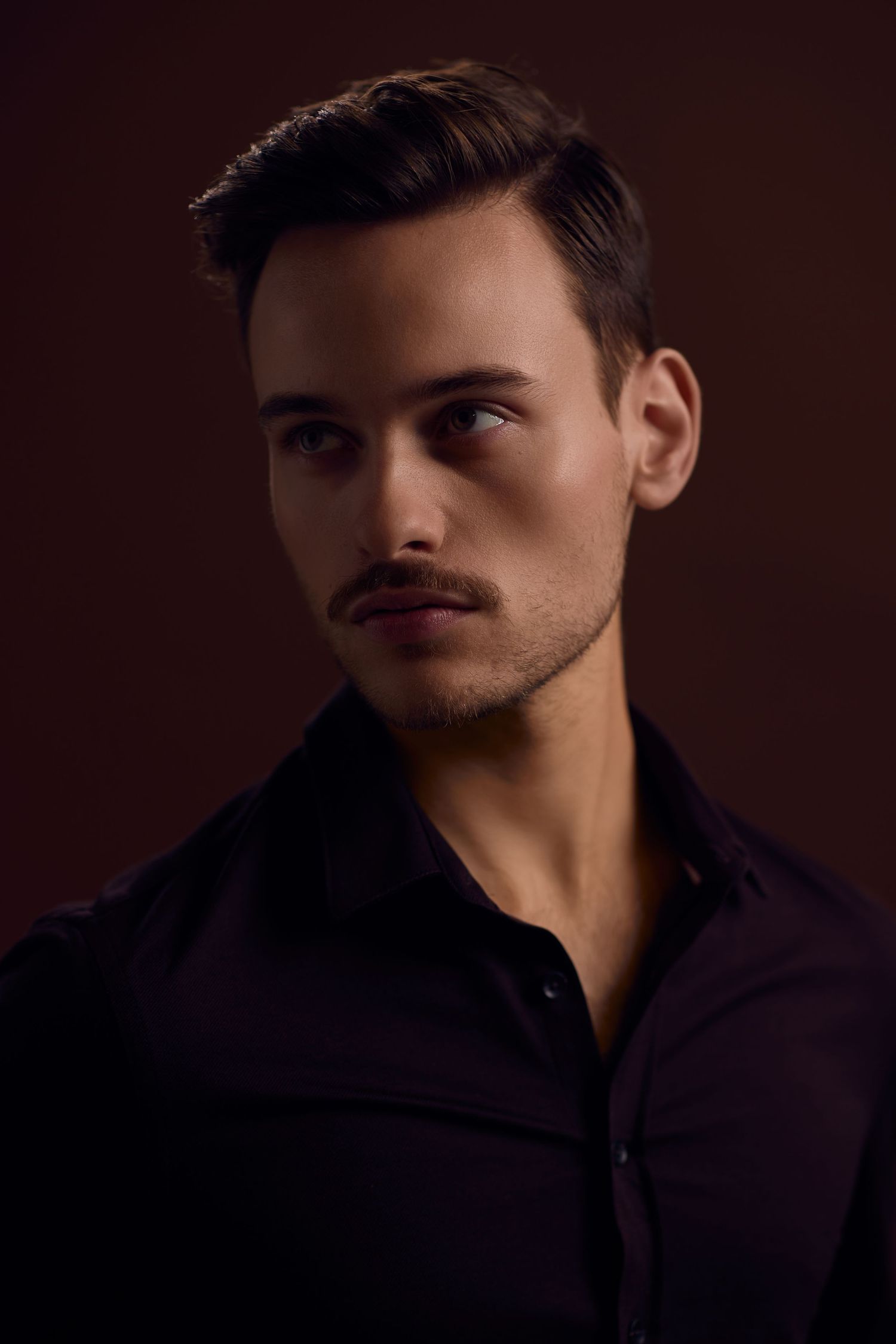 A dramatic side profile portrait in dark lighting shows a figure wearing a dark colored shirt against a deep brown background.