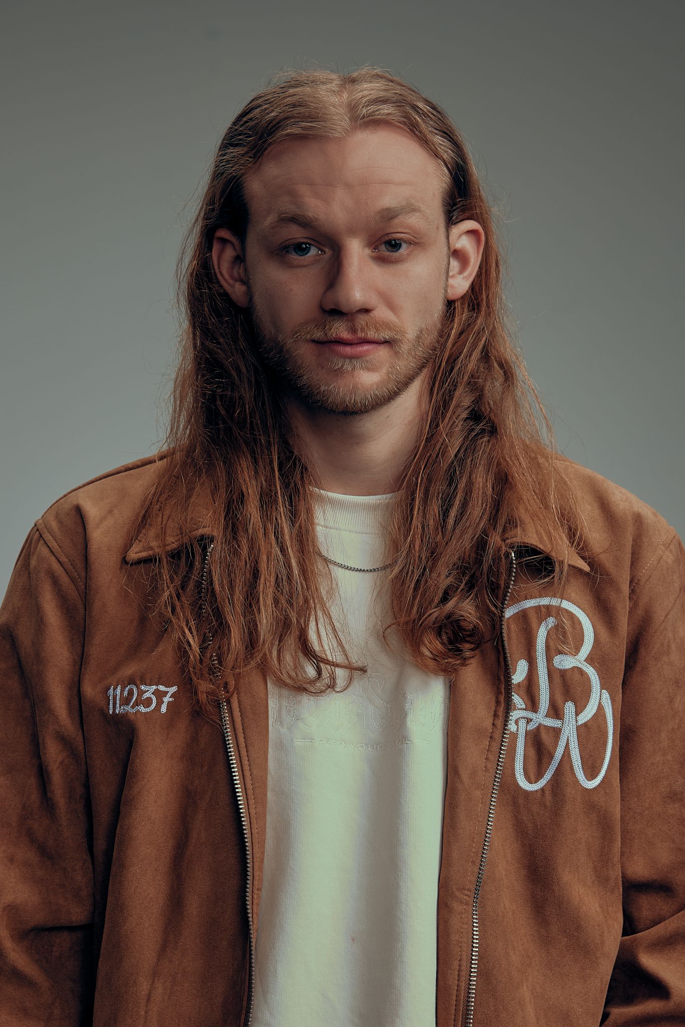 Portrait of long-haired person wearing brown suede jacket with graffiti numbers against gray wall.