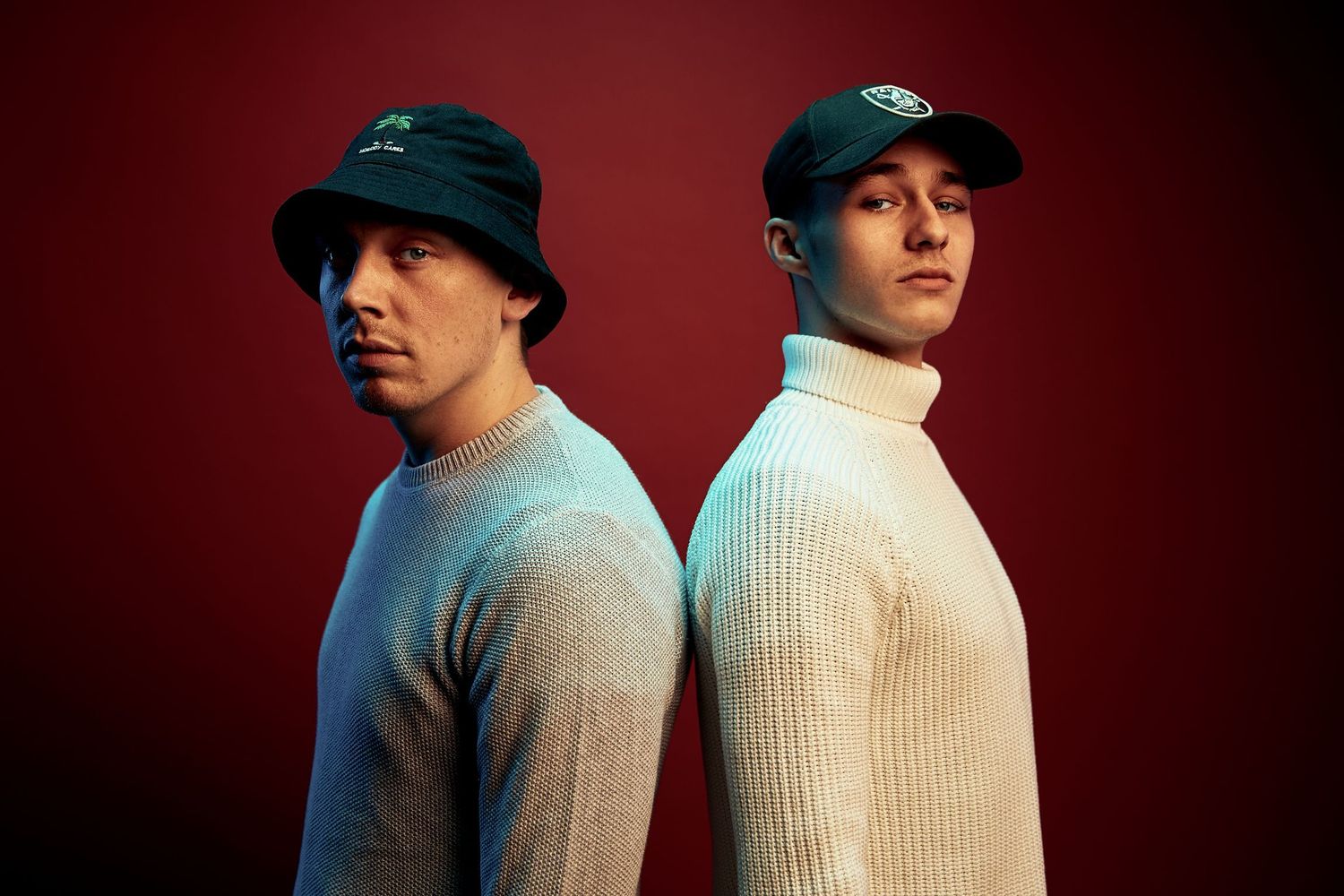 Two figures stand side by side in matching beige sweaters against a deep red background.