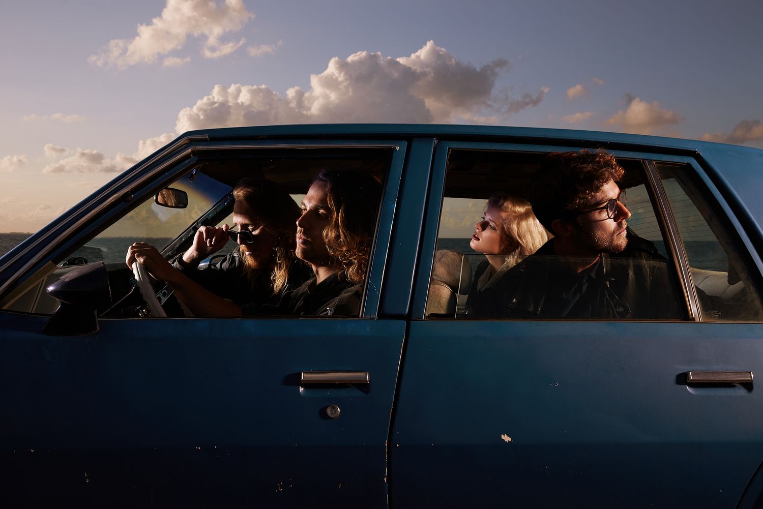 People sitting in blue car at sunset with dramatic sky.