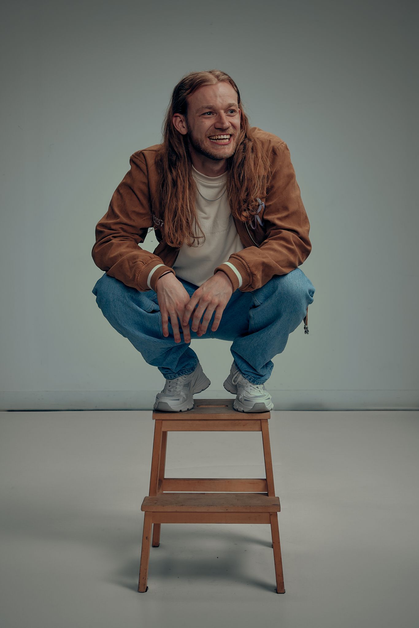 A person in a brown jacket and jeans crouches on a wooden step stool against a gray studio backdrop.