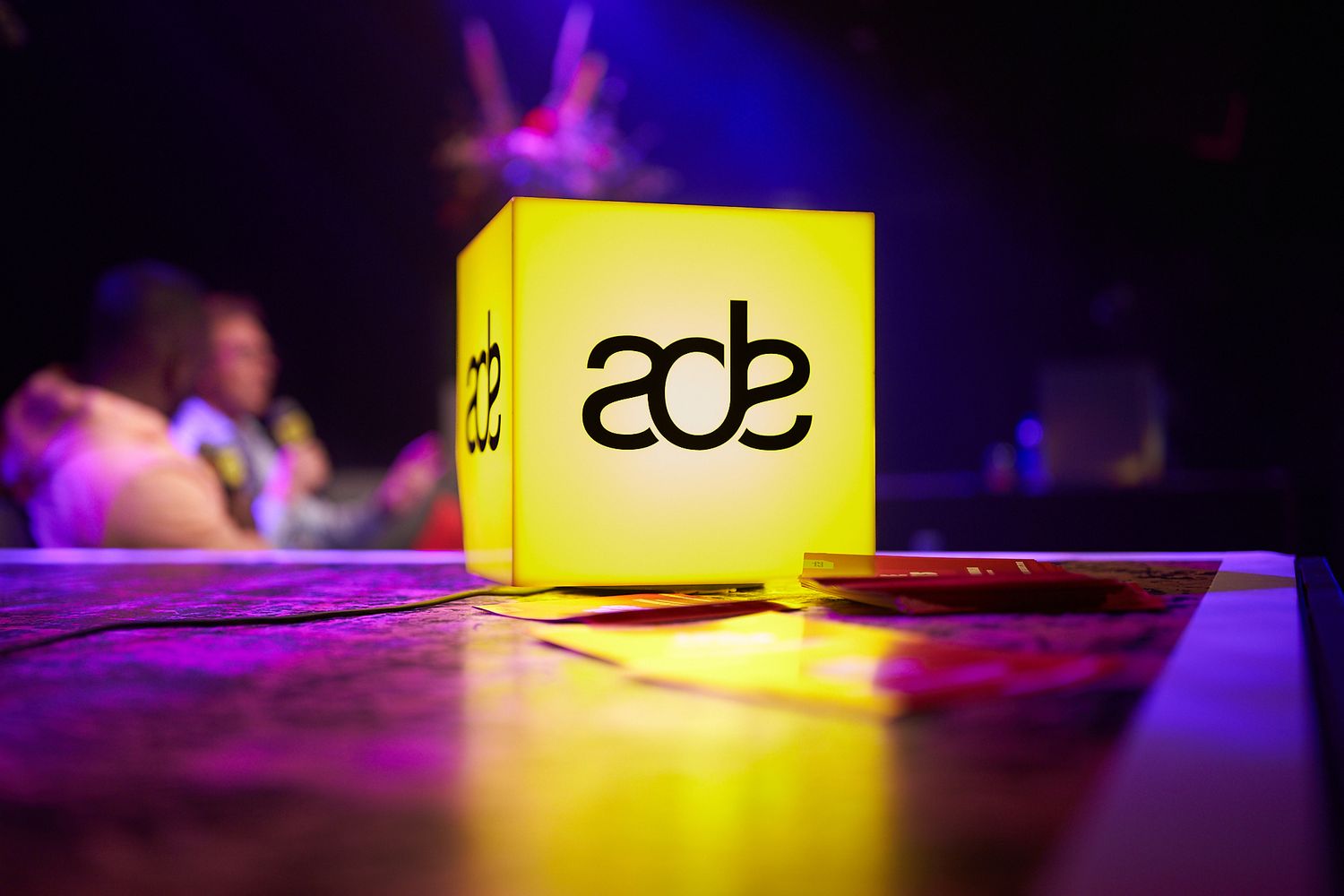 Illuminated yellow ADE sign glows in dark purple lighting at an event or club setting.