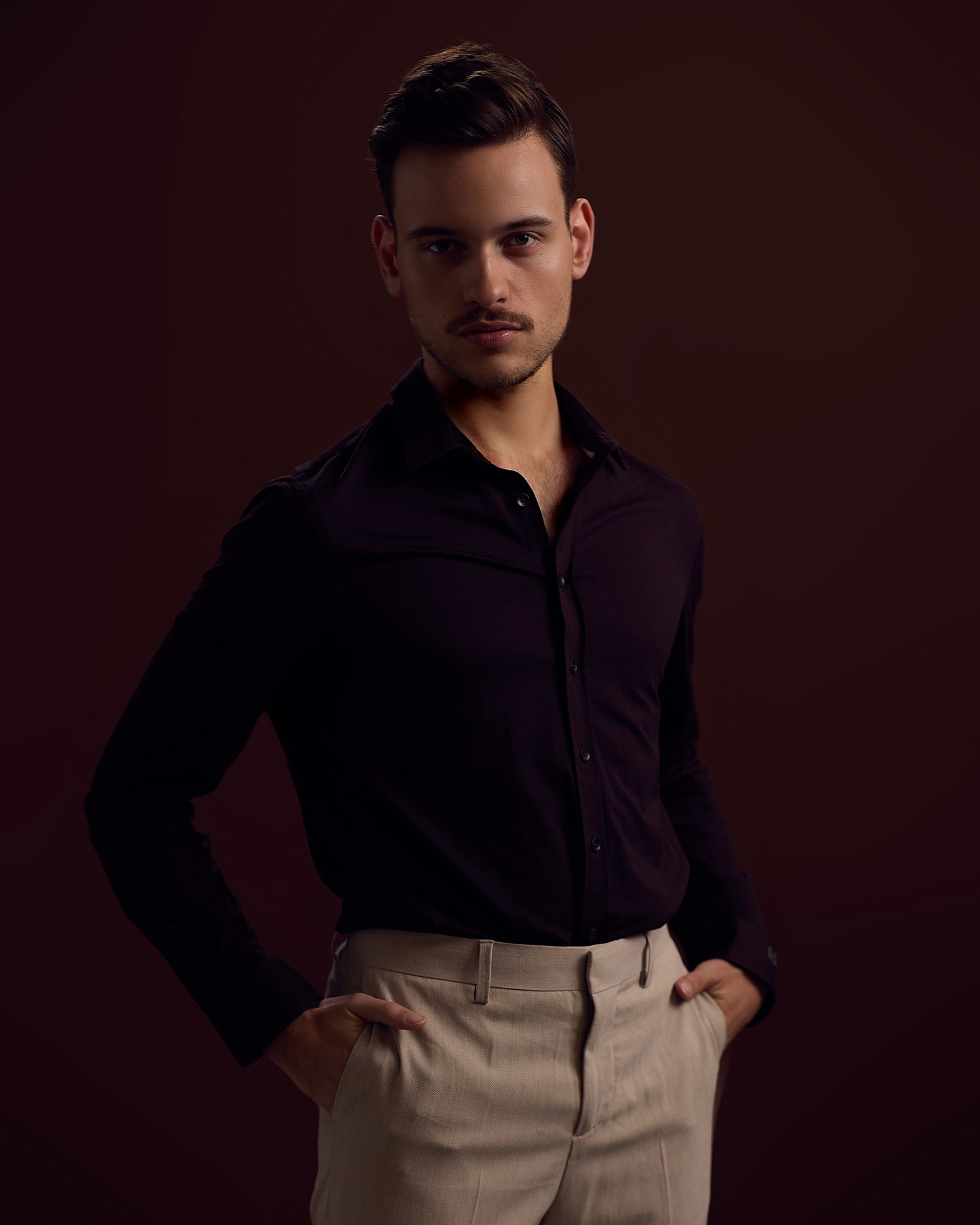 A model in a dark long-sleeve shirt and khaki pants poses against a dark background in a fashion photoshoot series.