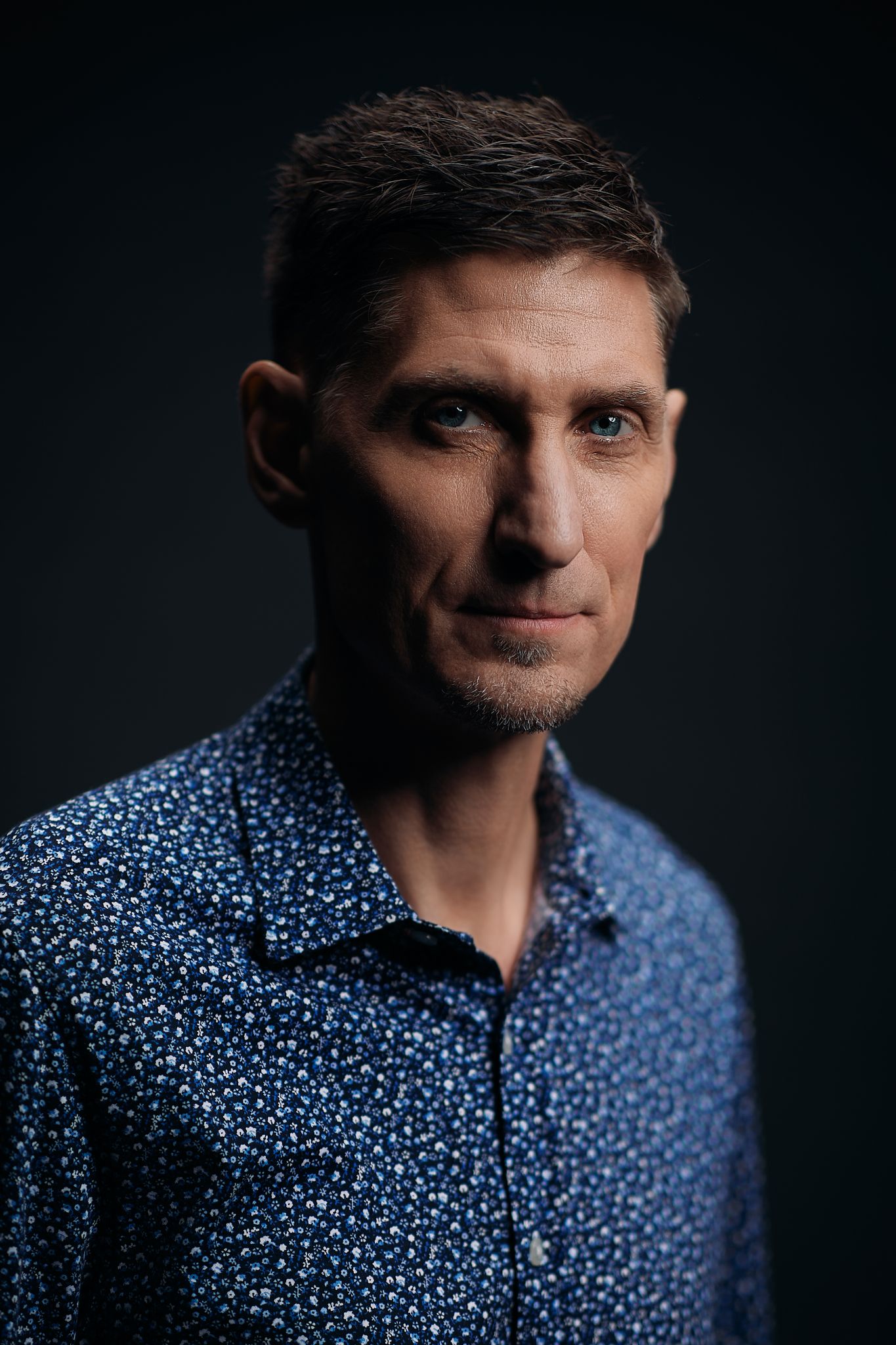 Portrait of person wearing patterned blue shirt against dark background.