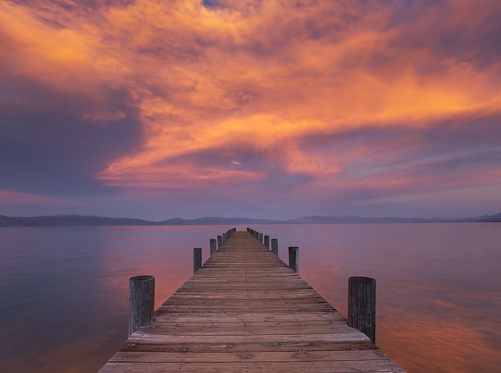 A photo of a wooden pier stretching into calm Lake Tahoe under dramatic purple and orange sunset clouds.