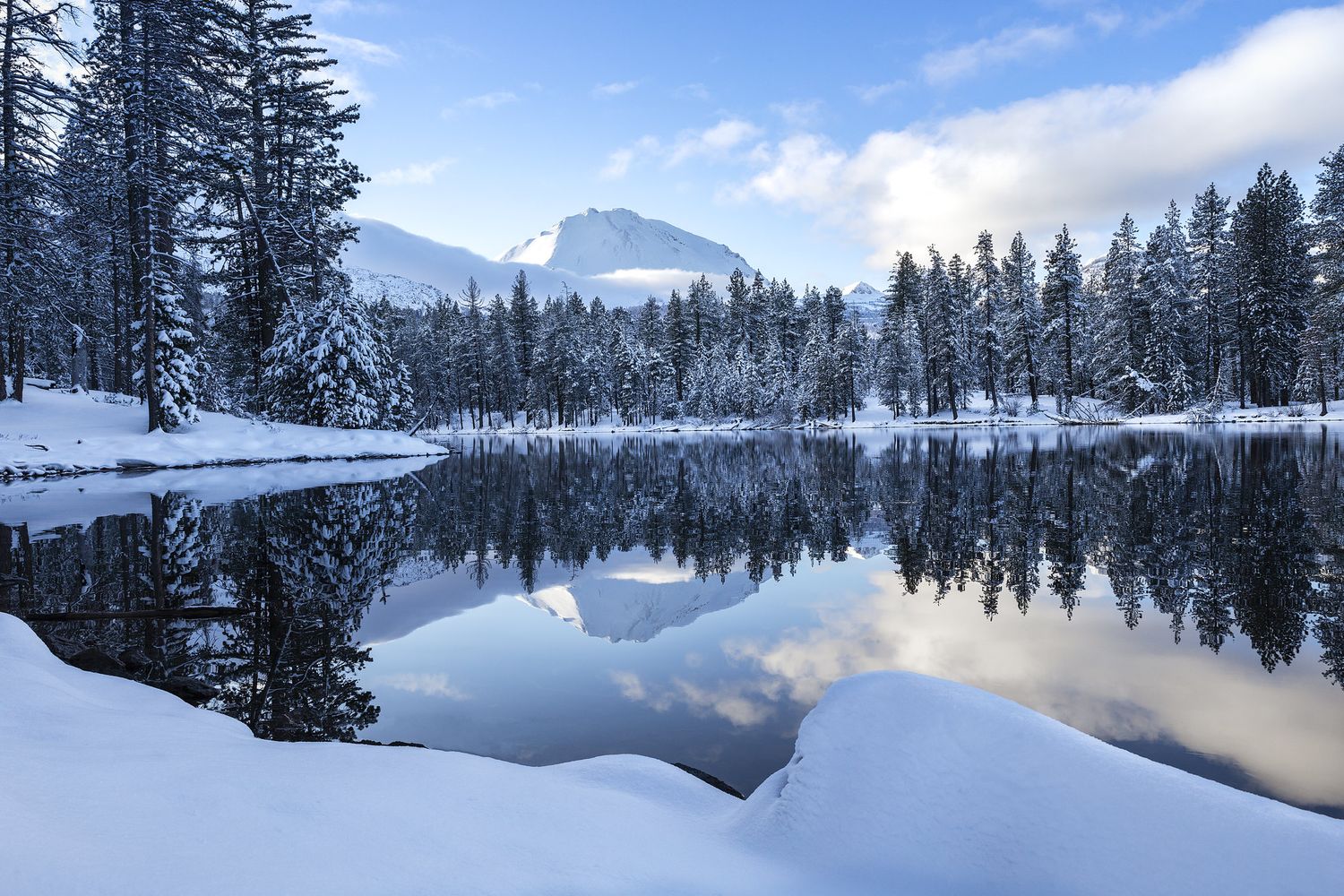 A photograph of snow-covered evergreen trees lining a tranquil mountain lake reflecting the cloudy winter sky and surrounding landscape.