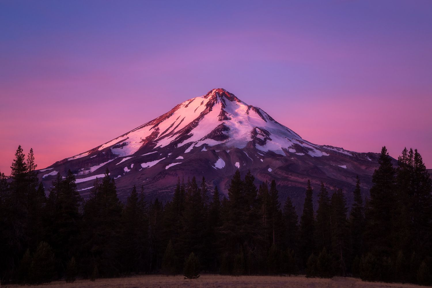 A photograph of scenic Mount Hood in Oregon at sunrise showing purple and pink sky colors over its snowy peak.
