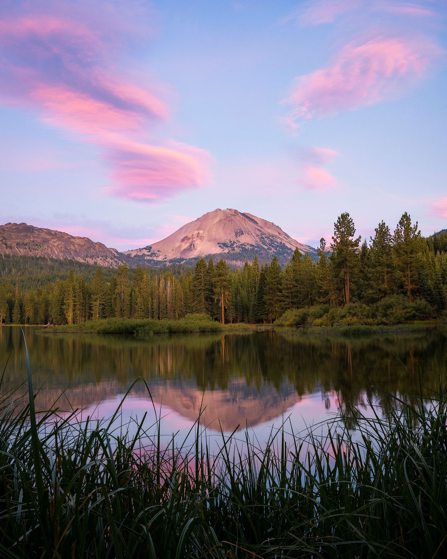 Serene mountain lake reflecting pink sunset clouds and a snow-capped volcanic peak surrounded by evergreen forest.