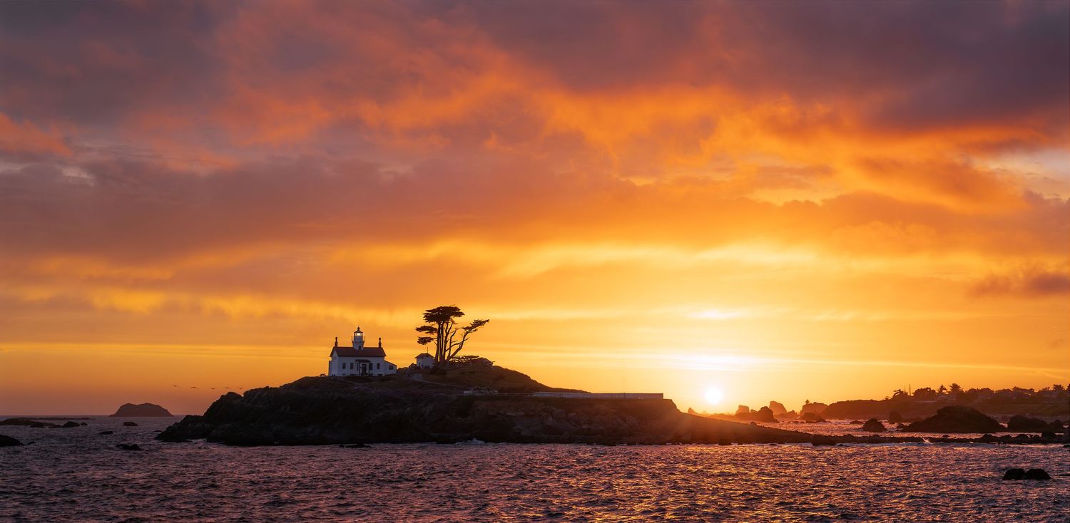 Stunning golden sunset over a rocky coastal island with a lighthouse and cypress tree silhouetted against vibrant orange skies.