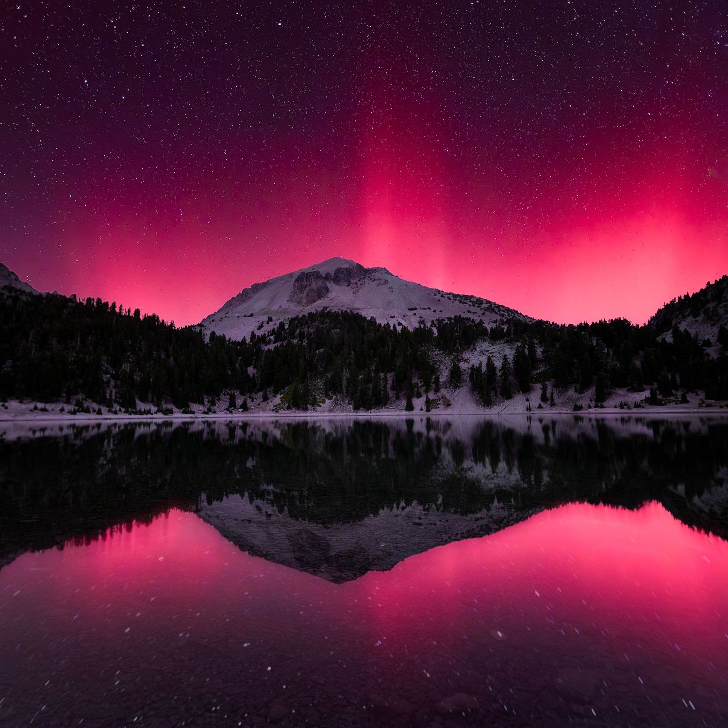 Snow-capped mountain reflects in calm lake under vibrant pink and red aurora borealis in starry night sky.