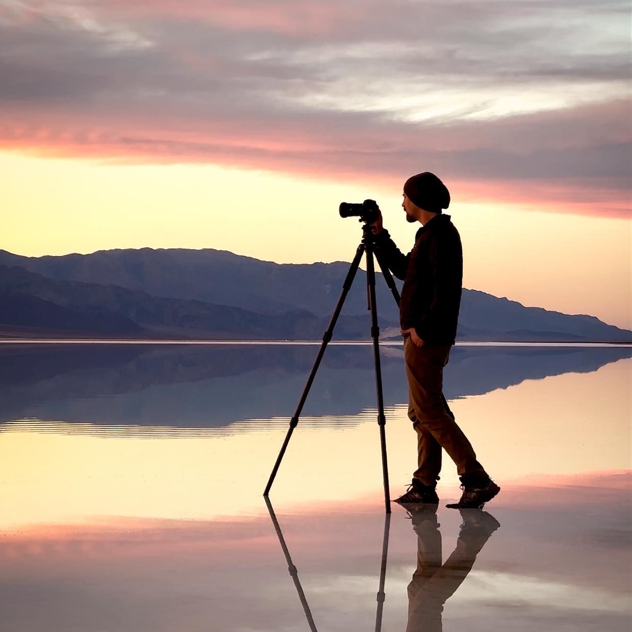 A silhouette of photographer Matt Haynie with a tripod capturing reflections at sunset with mountains in background.