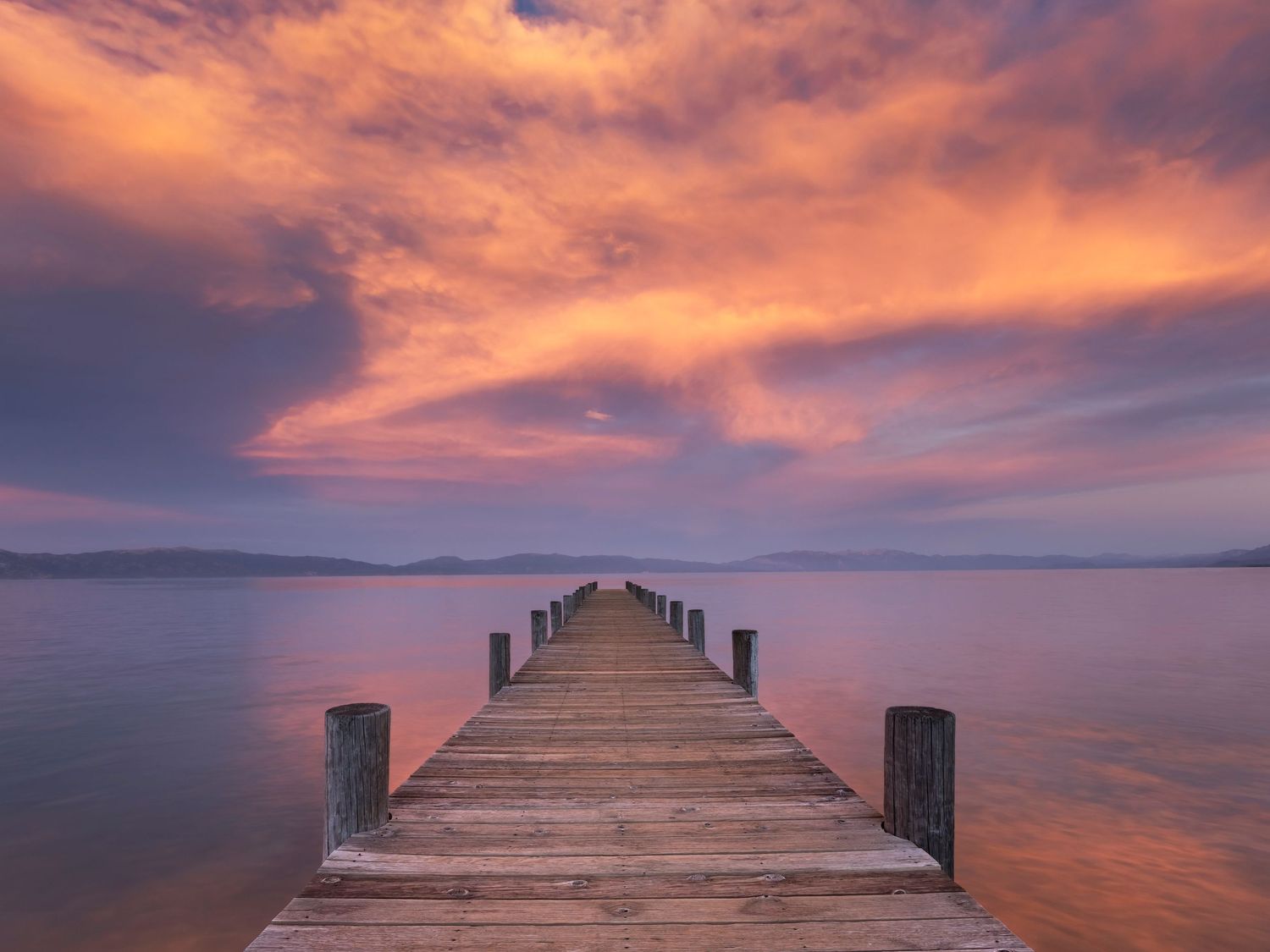 Wooden dock extending over calm Lake Tahoe at sunset with dramatic pink and orange clouds reflecting on still water.