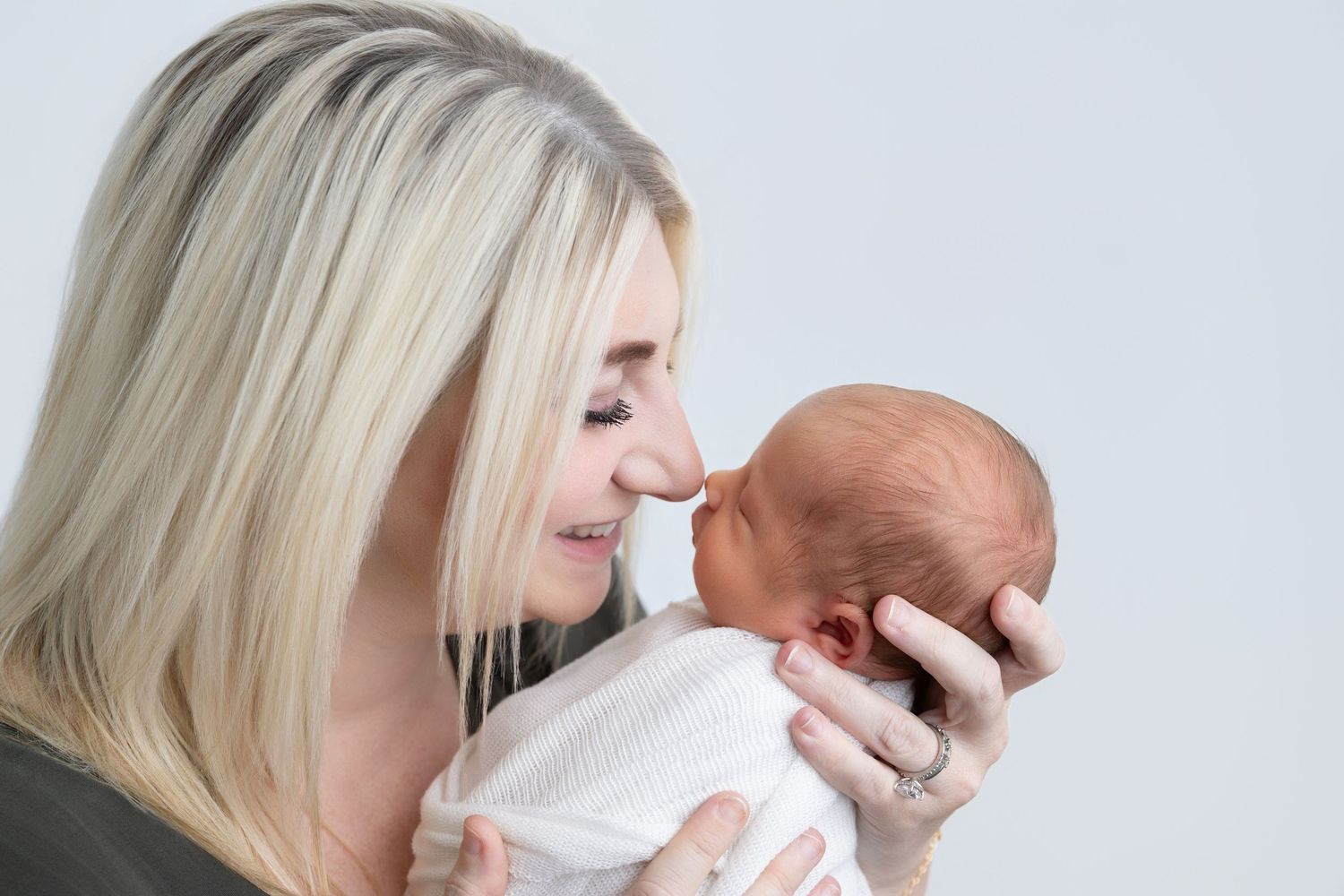 Blonde mother tenderly touching noses with her newborn baby wrapped in white, sharing a sweet bonding moment.